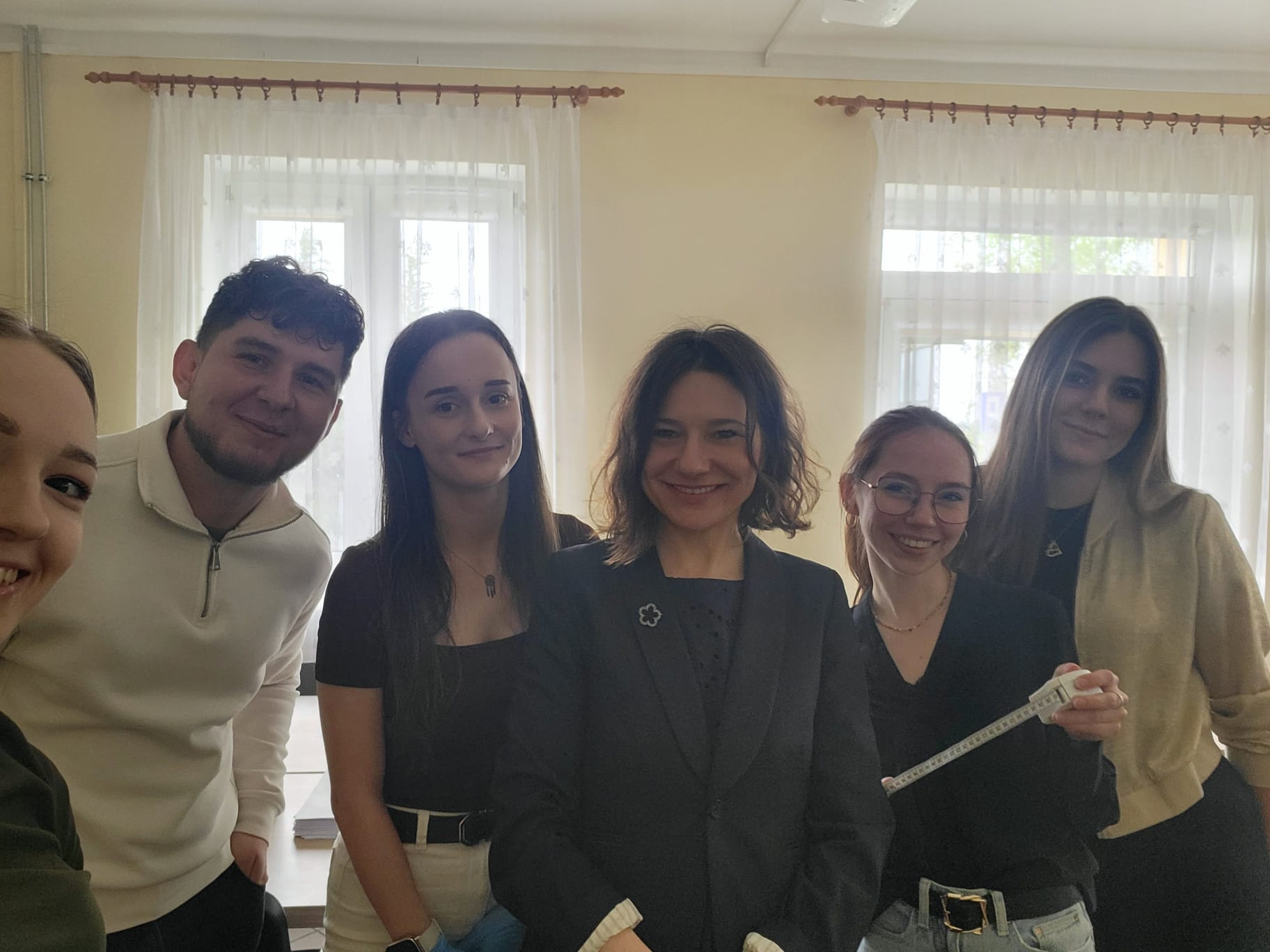Grupa uczestników stoi razem w jasnym pomieszczeniu i pozuje do wspólnego zdjęcia. Jedna z osób trzyma przyrząd pomiarowy, co sugeruje udział w zajęciach lub badaniach o charakterze edukacyjnym lub zdrowotnym. W tle widoczne są duże okna z jasnymi zasłonami.