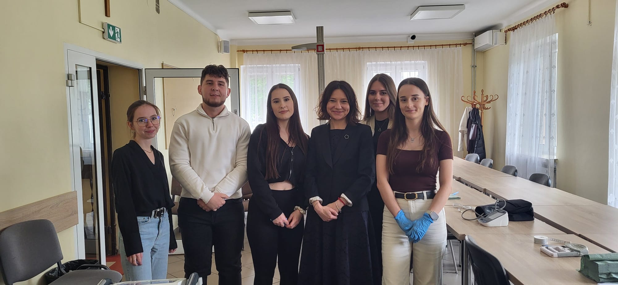Grupa uczestników stoi w jasnej sali i pozuje do wspólnego zdjęcia. W pomieszczeniu znajdują się stoły z wyposażeniem wykorzystywanym podczas badań lub zajęć, m.in. ciśnieniomierz i przyrządy pomiarowe. W tle widoczne są duże okna, krzesła oraz elementy wyposażenia sali o charakterze edukacyjnym lub medycznym.