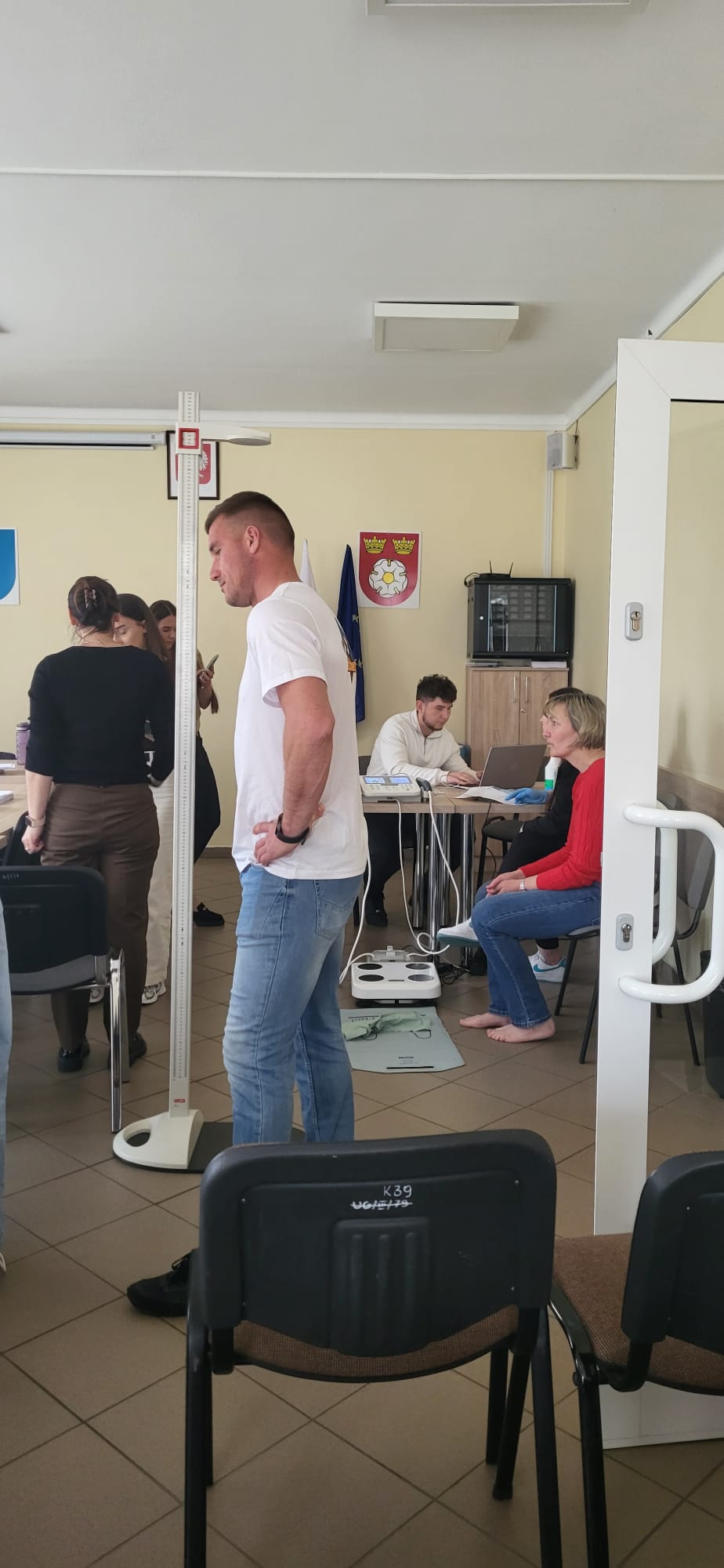 Uczestnicy biorą udział w badaniach lub zajęciach prowadzonych w sali o charakterze edukacyjnym lub medycznym. Jedna z osób stoi przy urządzeniu do pomiaru wzrostu, a w tle inni pracują przy stanowisku z laptopem i dokumentacją. W pomieszczeniu widoczne są krzesła, wagi elektroniczne oraz elementy dekoracyjne i flagi na ścianach.