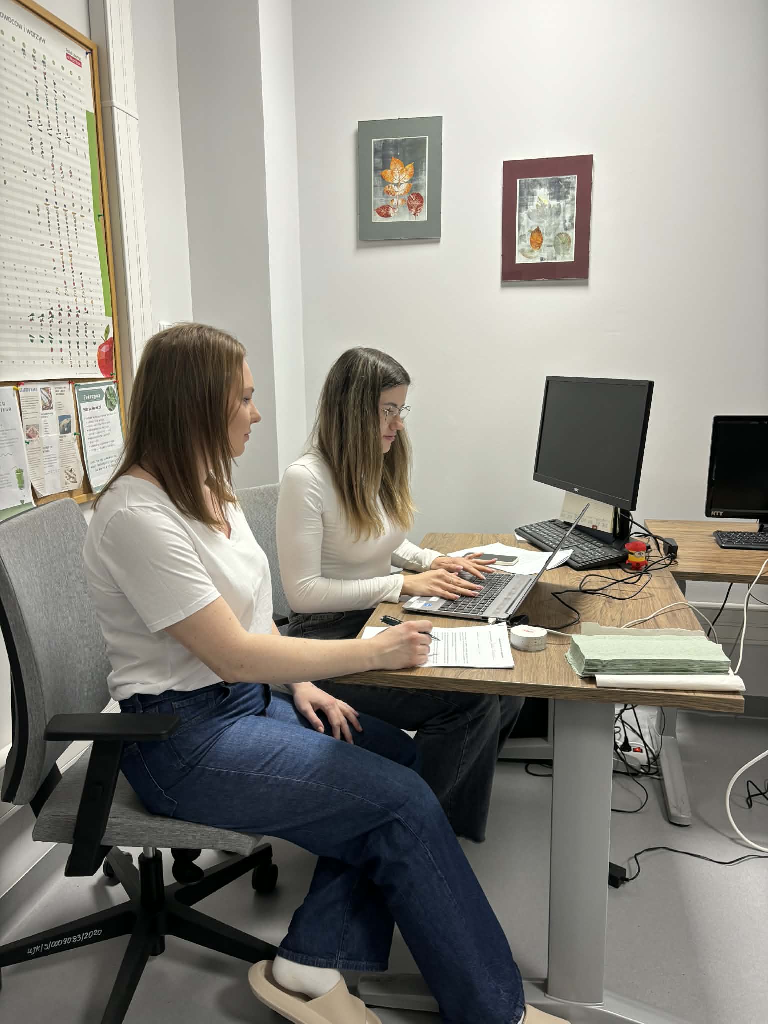 Dwie osoby pracują przy biurku w przestrzeni biurowej, zajmując się dokumentami i pracą na laptopie. Na stanowisku znajdują się monitory, dokumentacja oraz sprzęt elektroniczny. W tle widoczne są tablica z notatkami oraz oprawione grafiki, co podkreśla administracyjny lub organizacyjny charakter miejsca.