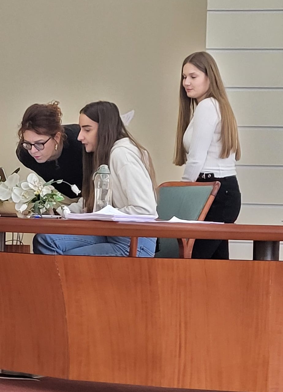 Trzy osoby pracują przy stole w sali o charakterze edukacyjnym lub organizacyjnym, skupiając się na ekranie laptopa. Na blacie znajdują się dokumenty, butelka wody oraz roślina doniczkowa. Jedna z osób stoi obok, obserwując przebieg pracy lub dyskusji.
