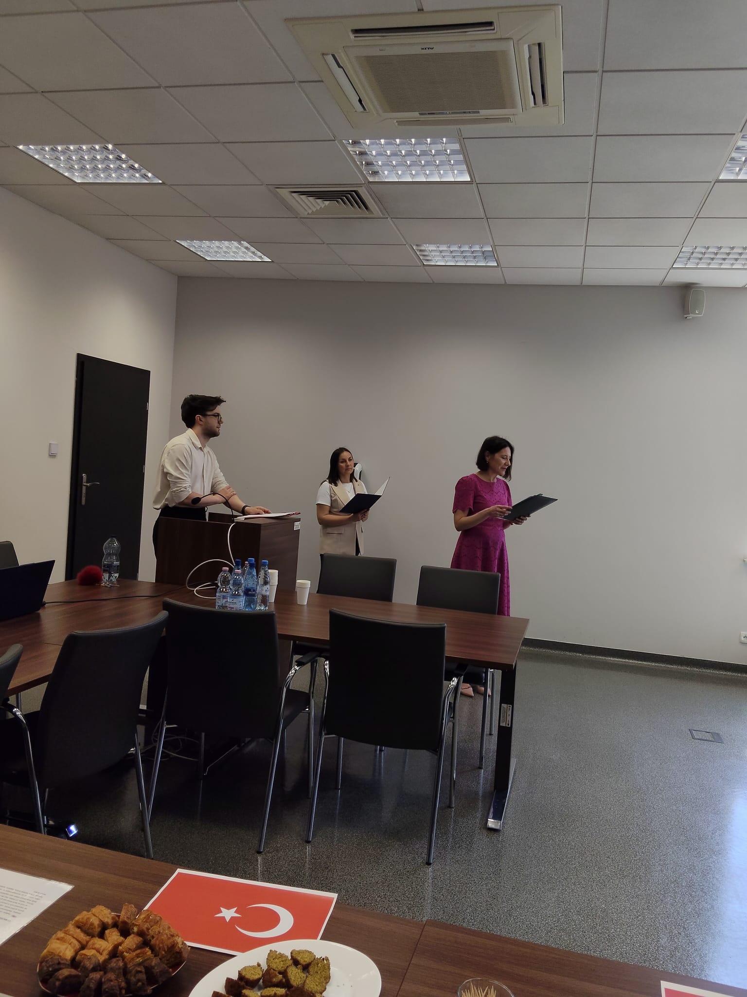 W sali konferencyjnej trzy osoby stoją przy stole i trzymają otwarte teczki lub dokumenty, przygotowując prezentację lub wystąpienie. Na stole znajdują się materiały, laptop i butelki z wodą, a w tle widoczne są krzesła konferencyjne i proste wyposażenie sali. Na pierwszym planie ustawiono poczęstunek oraz elementy związane z prezentacją kuchni lub kultury tureckiej.
