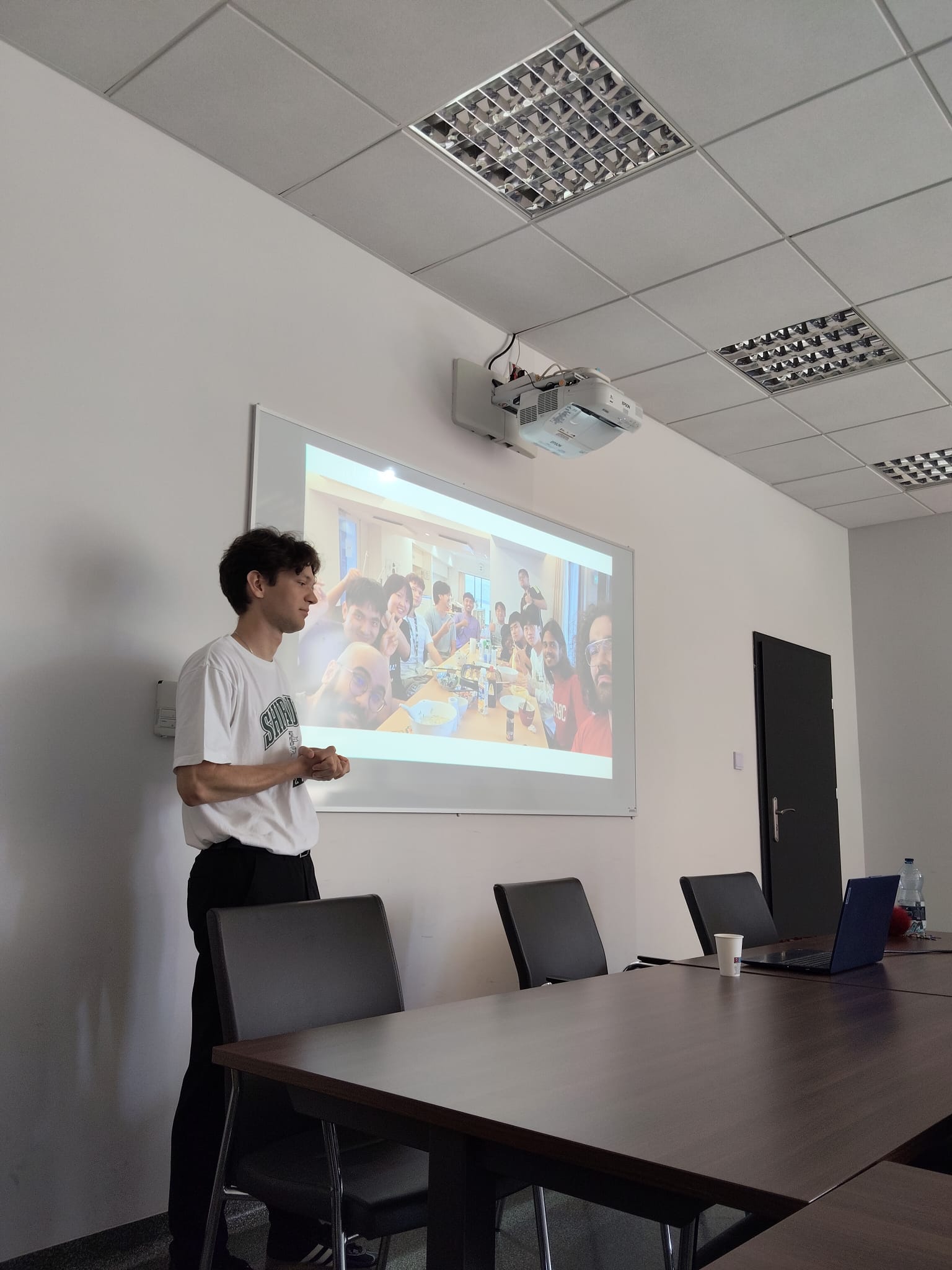 Osoba prowadzi prezentację w nowoczesnej sali konferencyjnej, stojąc obok ekranu z wyświetlonym zdjęciem przedstawiającym spotkanie przy wspólnym stole. W pomieszczeniu znajdują się stół konferencyjny, krzesła, laptop oraz tablica, co podkreśla szkoleniowy lub warsztatowy charakter wydarzenia.