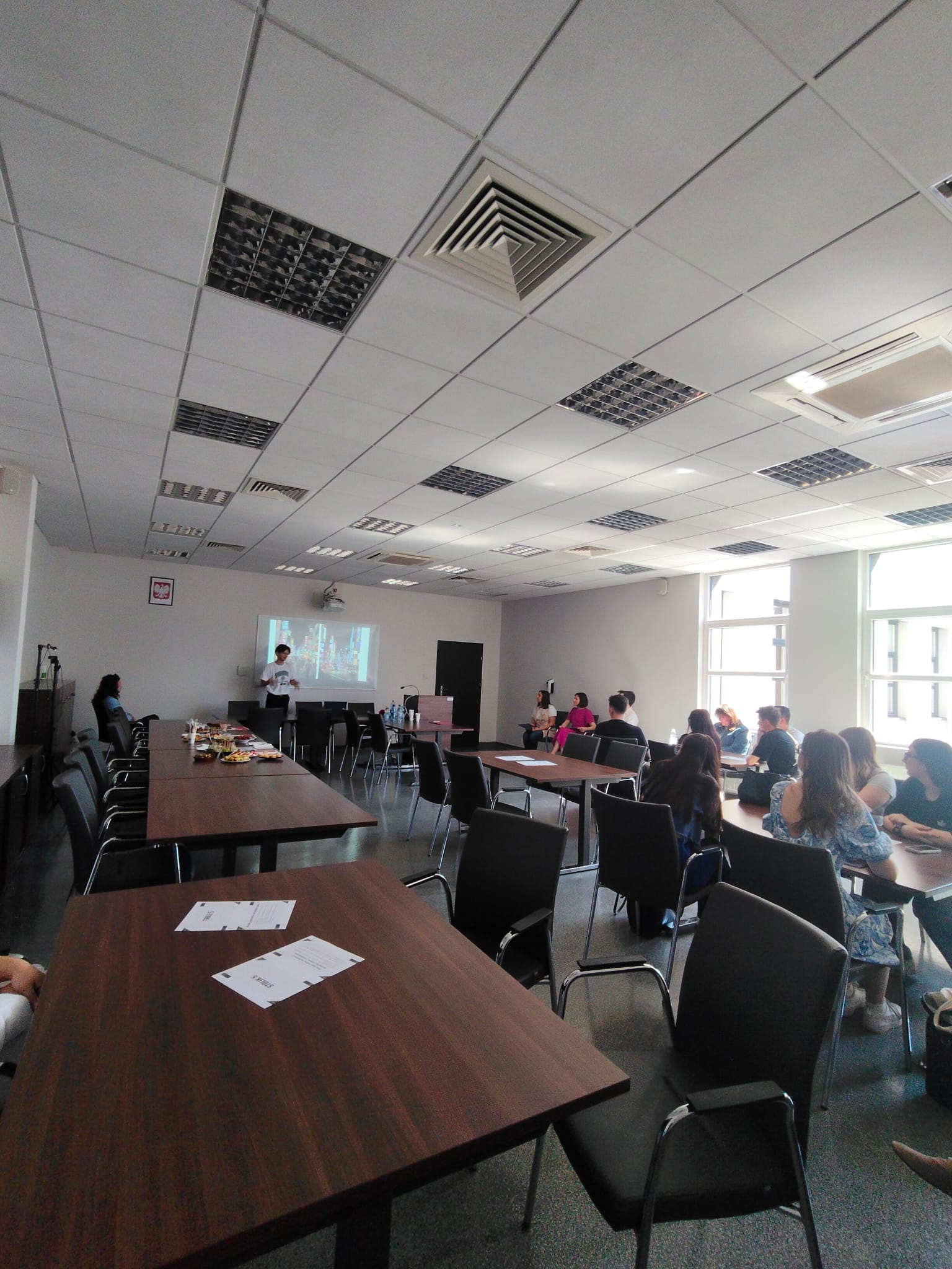 W przestronnej sali konferencyjnej odbywa się spotkanie lub warsztat z prezentacją wyświetlaną na ekranie. Uczestnicy siedzą przy stołach ustawionych w różnych częściach pomieszczenia i słuchają osoby prowadzącej. W tle widoczne są duże okna zapewniające naturalne światło oraz stół z przekąskami i napojami.