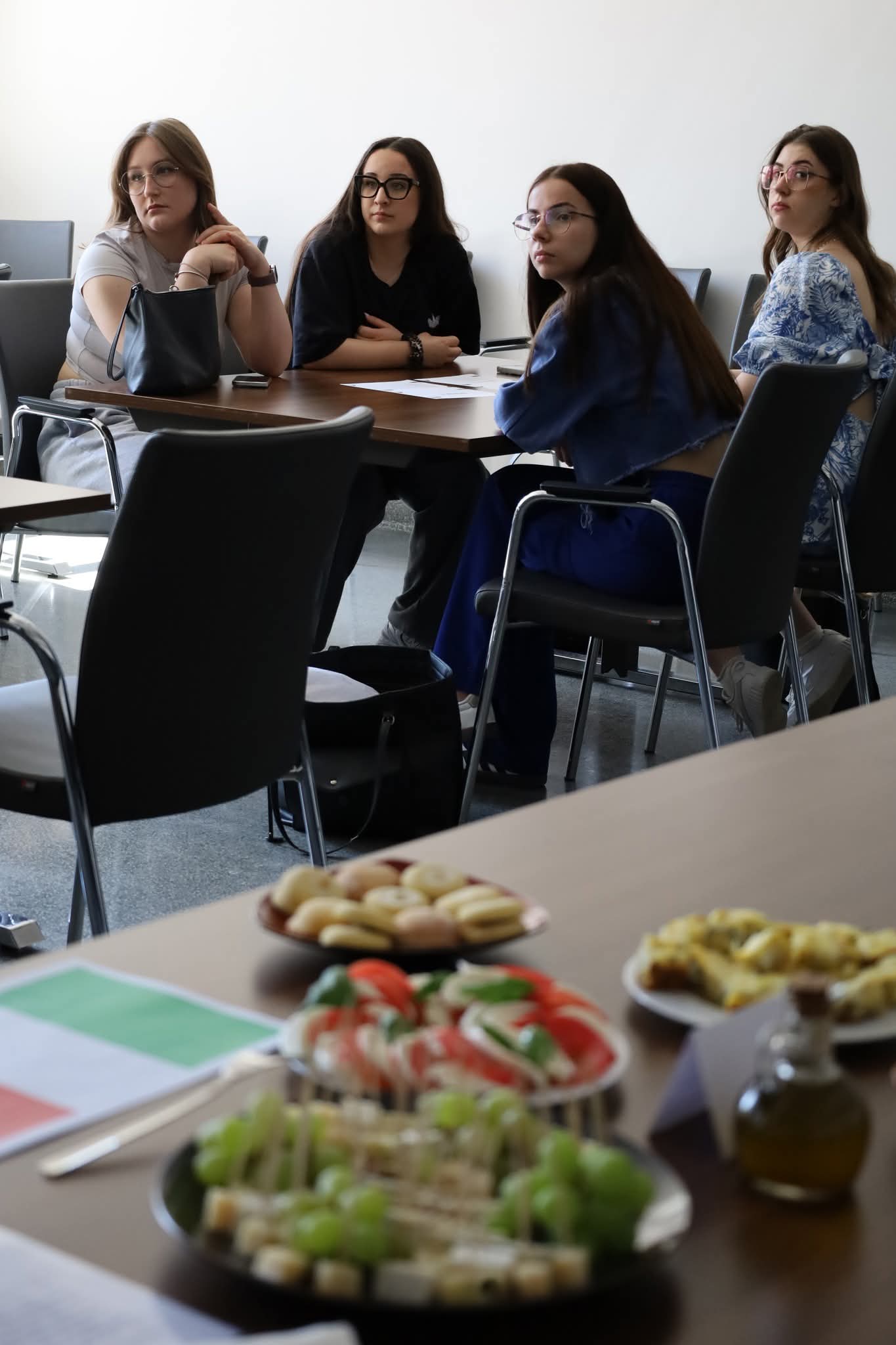 Uczestnicy siedzą przy stole konferencyjnym podczas spotkania lub warsztatów, skupiając się na rozmowie i prezentowanych treściach. Na pierwszym planie widoczny jest poczęstunek z przekąskami, owocami i warzywami, co nadaje spotkaniu swobodną i integracyjną atmosferę.