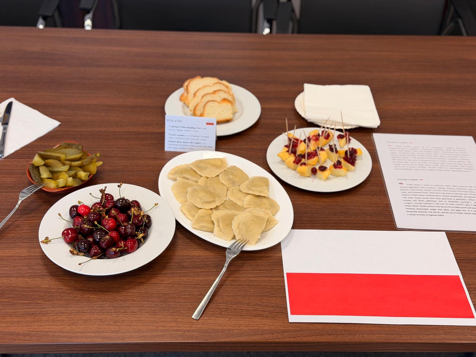 Na drewnianym stole ustawiono różne przekąski i potrawy, w tym pierogi, owoce, pieczywo oraz małe przekąski na wykałaczkach. Obok znajdują się dodatki, serwetki i materiały związane z wydarzeniem, co sugeruje poczęstunek podczas spotkania lub warsztatów.