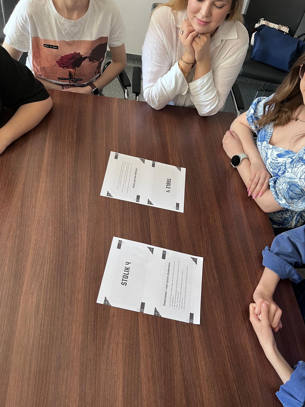 Pięć osób siedzi przy prostokątnym stole podczas spotkania lub wydarzenia organizacyjnego. Na środku stołu znajdują się kartki z oznaczeniem „Stolik 4 / Table 4”, wskazujące numer miejsca lub grupy. Widoczne są jedynie fragmenty uczestników oraz proste otoczenie sali.