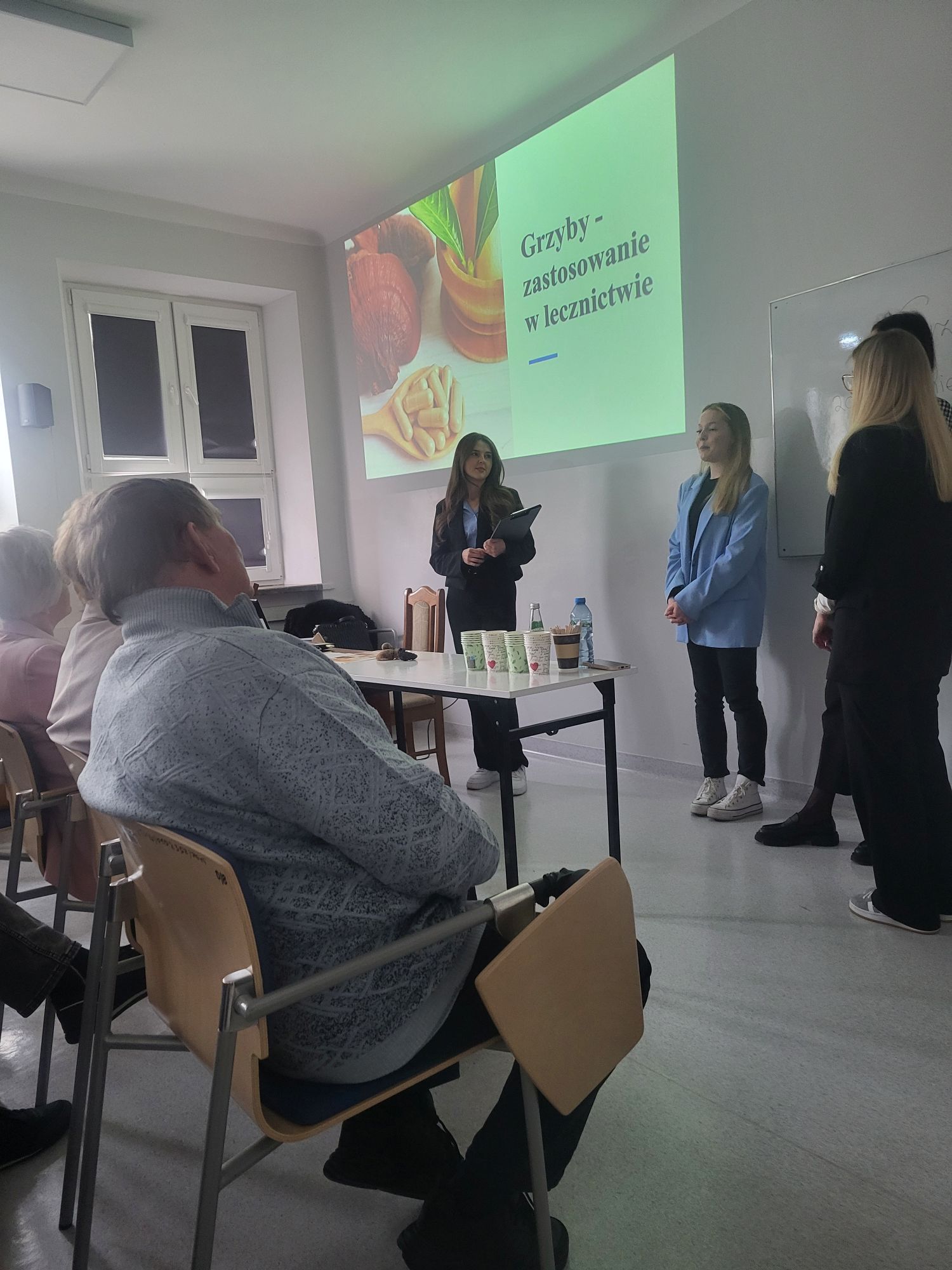 W sali dydaktycznej odbywa się prezentacja prowadzona przez zespół prelegentek. Jedna z osób przemawia do zgromadzonych uczestników, podczas gdy pozostałe wspierają wystąpienie. Publiczność siedzi w rzędach i uważnie słucha prezentacji wyświetlanej na ekranie, a przygotowane na stole materiały i napoje podkreślają warsztatowy charakter wydarzenia.