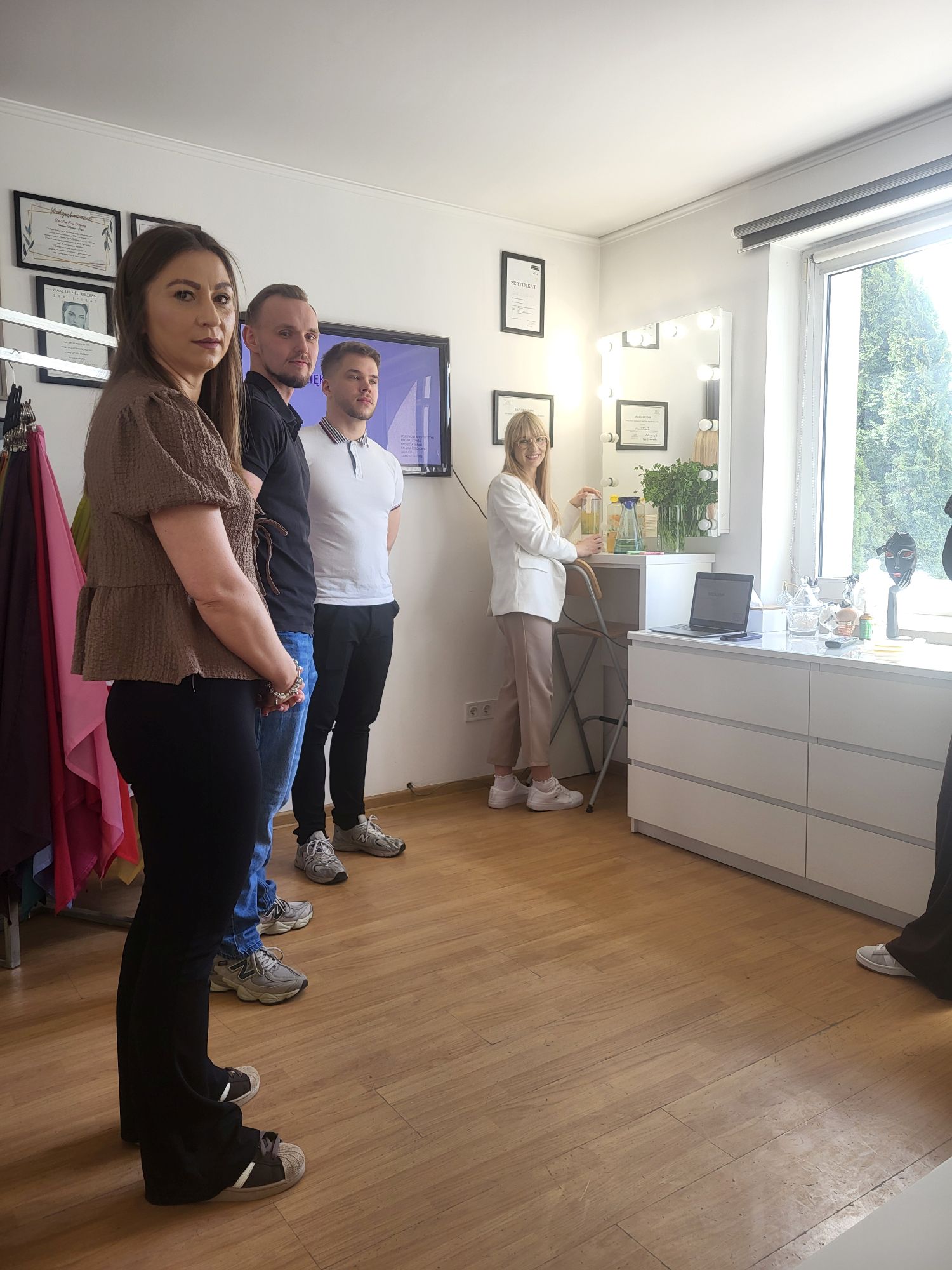 Grupa uczestników znajduje się w jasnym pomieszczeniu o charakterze dydaktycznym lub warsztatowym. Osoby stoją obok siebie i słuchają prezentacji lub instruktażu prowadzonego poza kadrem. Wnętrze wyposażone jest w materiały edukacyjne, certyfikaty na ścianach oraz stanowisko z lustrem i oświetleniem, co sugeruje wykorzystanie sali do zajęć praktycznych lub szkoleniowych.