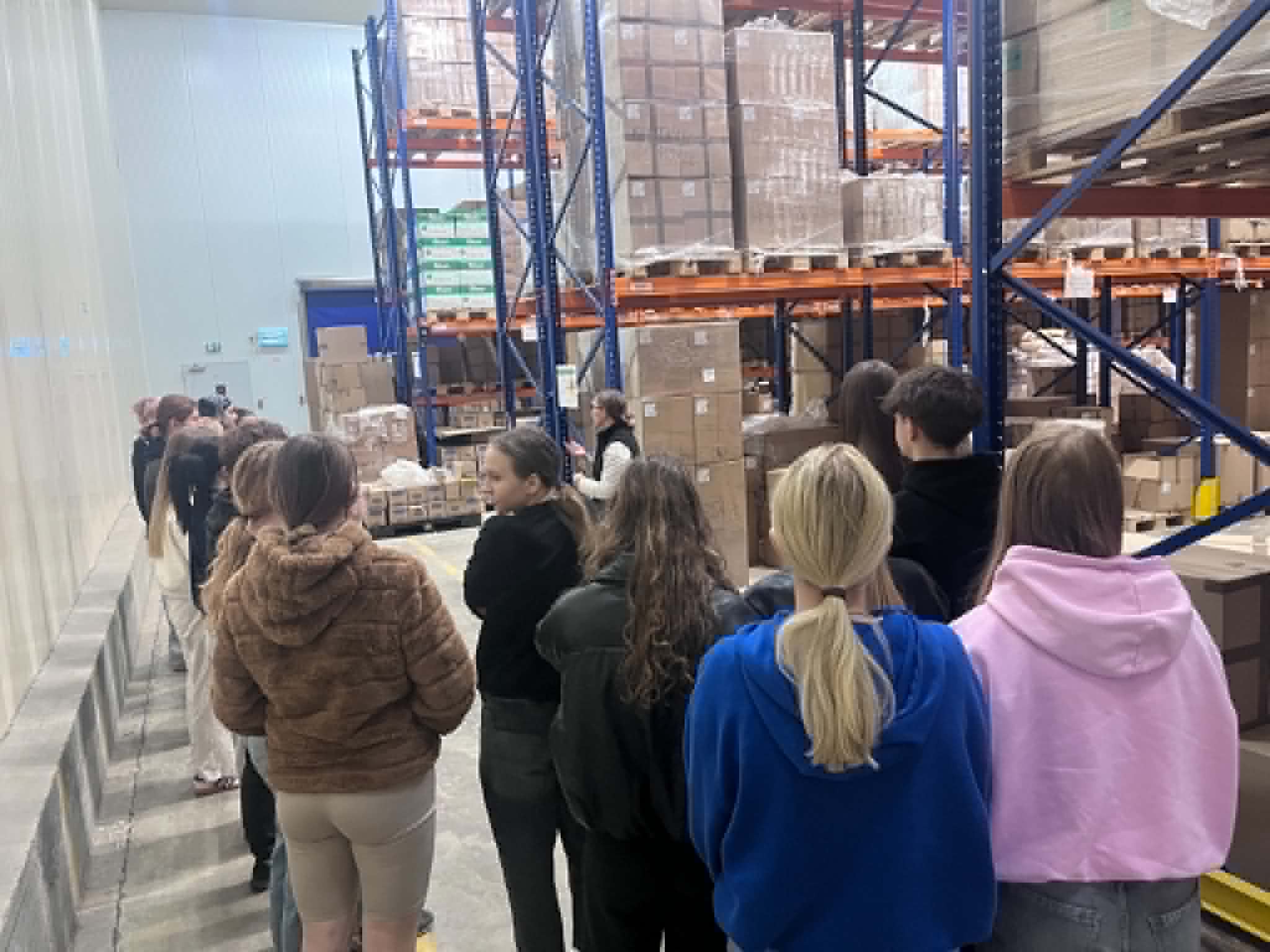 Grupa uczestników stoi w kolejce w przestrzeni magazynowej, prawdopodobnie podczas wizyty studyjnej, szkolenia lub zwiedzania obiektu. W tle widoczne są wysokie regały magazynowe wypełnione kartonami i zapakowanymi produktami, charakterystyczne dla nowoczesnego centrum logistycznego. Uczestnicy słuchają instrukcji lub oczekują na kolejny etap prezentacji dotyczącej funkcjonowania magazynu.