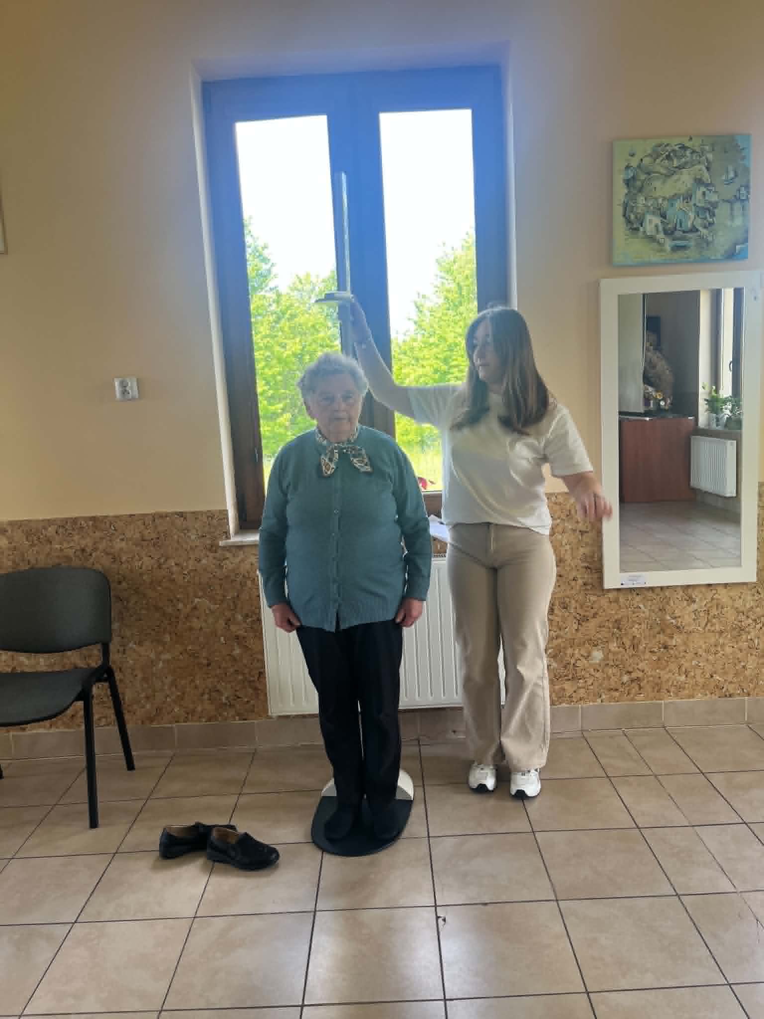 Uczestniczka bierze udział w badaniu lub konsultacji zdrowotnej przy wykorzystaniu specjalistycznego urządzenia pomiarowego. Obok znajduje się osoba nadzorująca przebieg pomiaru, natomiast w tle widoczna jest osoba dokumentująca wyniki na laptopie i w formularzach. Całość odbywa się w pomieszczeniu o charakterze profilaktycznym lub diagnostycznym.