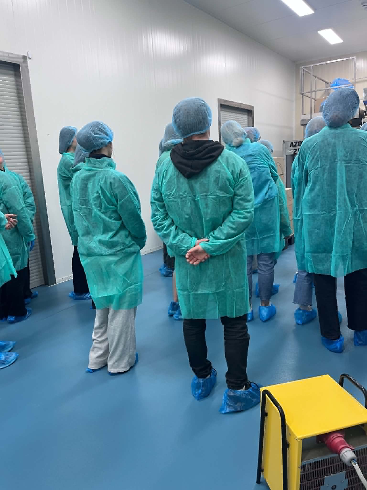 Grupa uczestników znajduje się w strefie o podwyższonych wymaganiach higienicznych, przygotowana do wizyty lub szkolenia w zakładzie produkcyjnym, laboratorium bądź pomieszczeniu typu cleanroom. Wszyscy ubrani są w jednorazowe fartuchy ochronne, czepki oraz ochraniacze na obuwie, co zapewnia zachowanie odpowiednich standardów czystości. W tle widoczne są elementy wyposażenia technicznego oraz infrastruktura charakterystyczna dla środowiska przemysłowego lub laboratoryjnego.