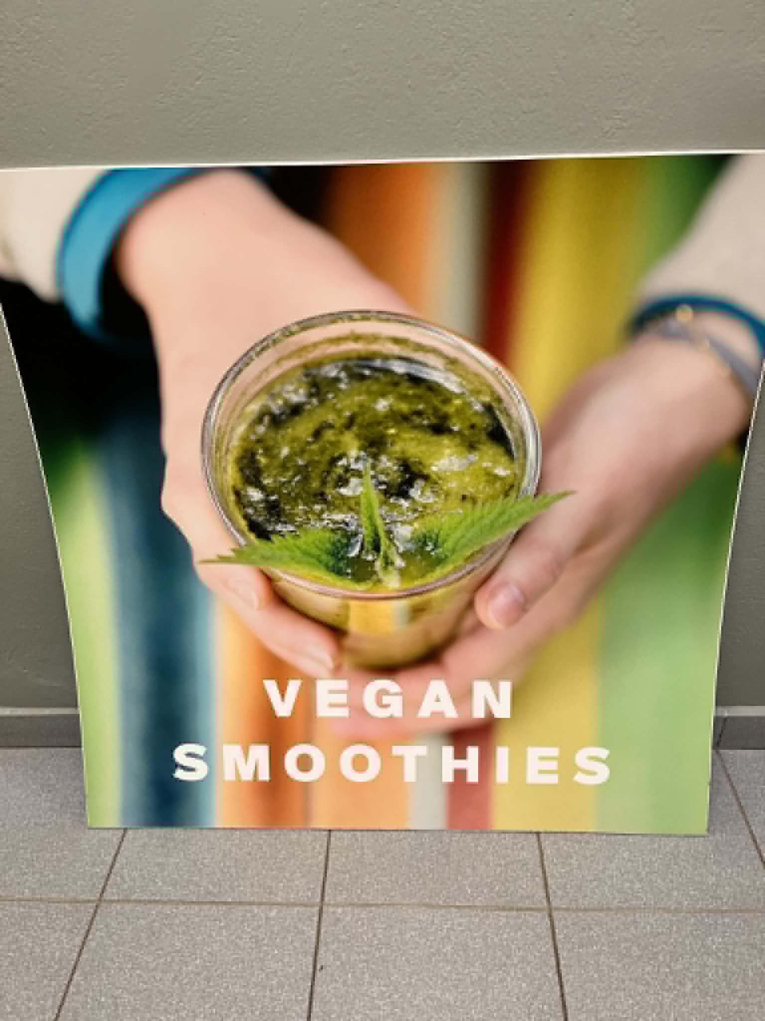 Plakat promocyjny przedstawiający szklankę zielonego smoothie trzymaną w dłoniach. Kolorowe tło oraz wyraźny napis „VEGAN SMOOTHIES” podkreślają tematykę zdrowego odżywiania i napojów roślinnych. Grafika może stanowić element promocji warsztatów, wydarzenia kulinarnego lub kampanii związanej z dietą wegańską i zdrowym stylem życia.