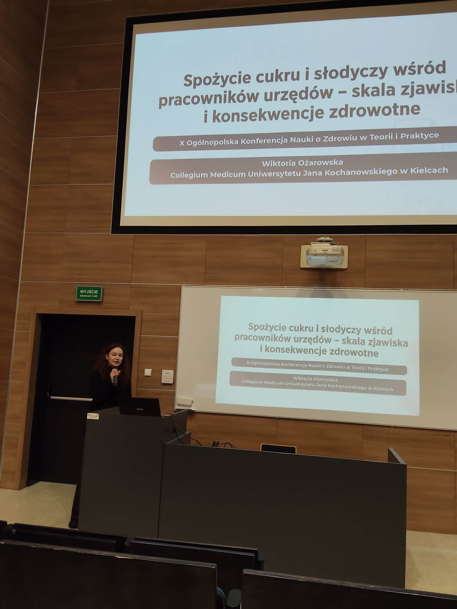 W auli wykładowej trwa prezentacja naukowa prowadzona przez prelegentkę stojącą przy mównicy z mikrofonem. Treść wystąpienia wyświetlana jest jednocześnie na dwóch ekranach projekcyjnych, co ułatwia odbiór prezentowanych materiałów. Nowoczesne wyposażenie audiowizualne oraz układ sali podkreślają konferencyjny i akademicki charakter wydarzenia.