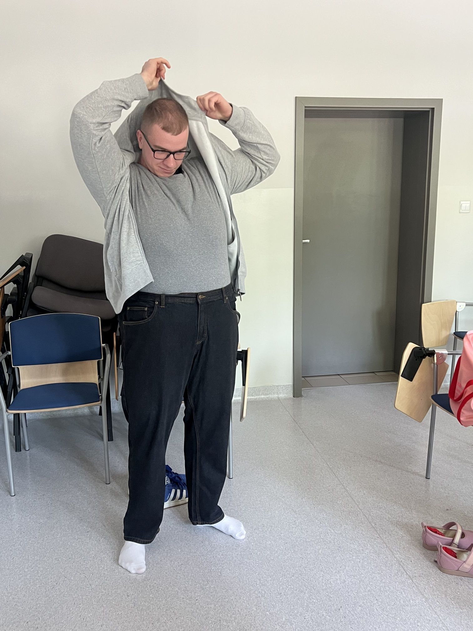 Osoba przygotowuje się do zajęć praktycznych lub ćwiczenia, zakładając dodatkowy element odzieży w sali szkoleniowej. W pomieszczeniu widoczne są krzesła, torby oraz pozostawione obuwie, co wskazuje na trwające warsztaty lub zajęcia grupowe. Jasne wnętrze i proste wyposażenie podkreślają edukacyjny charakter miejsca.