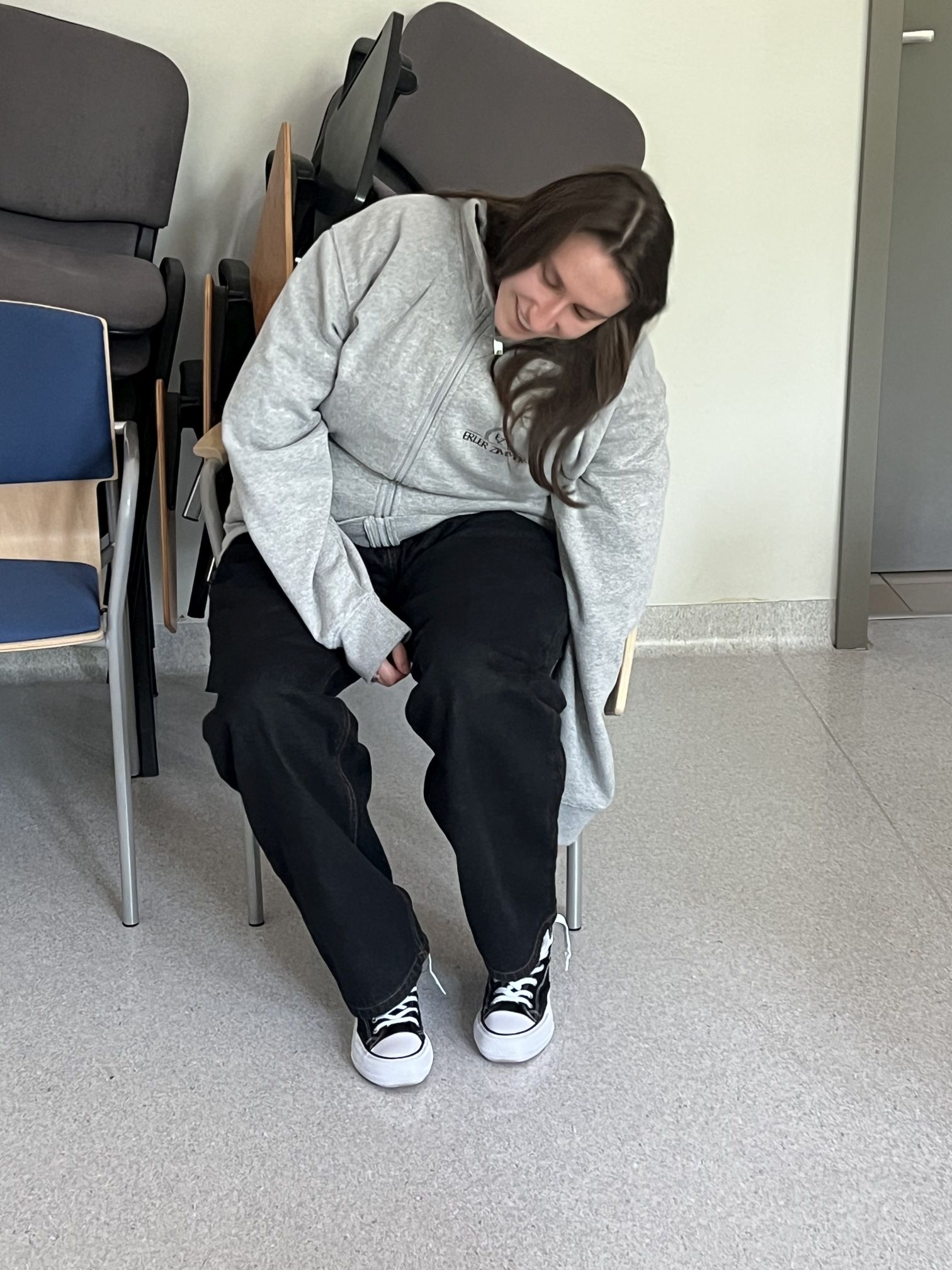 Osoba siedzi na krześle w sali dydaktycznej lub szkoleniowej, oczekując na rozpoczęcie zajęć lub uczestnicząc w ćwiczeniu. W tle widoczne są ustawione pod ścianą krzesła, a proste wyposażenie pomieszczenia podkreśla jego funkcjonalny charakter.
