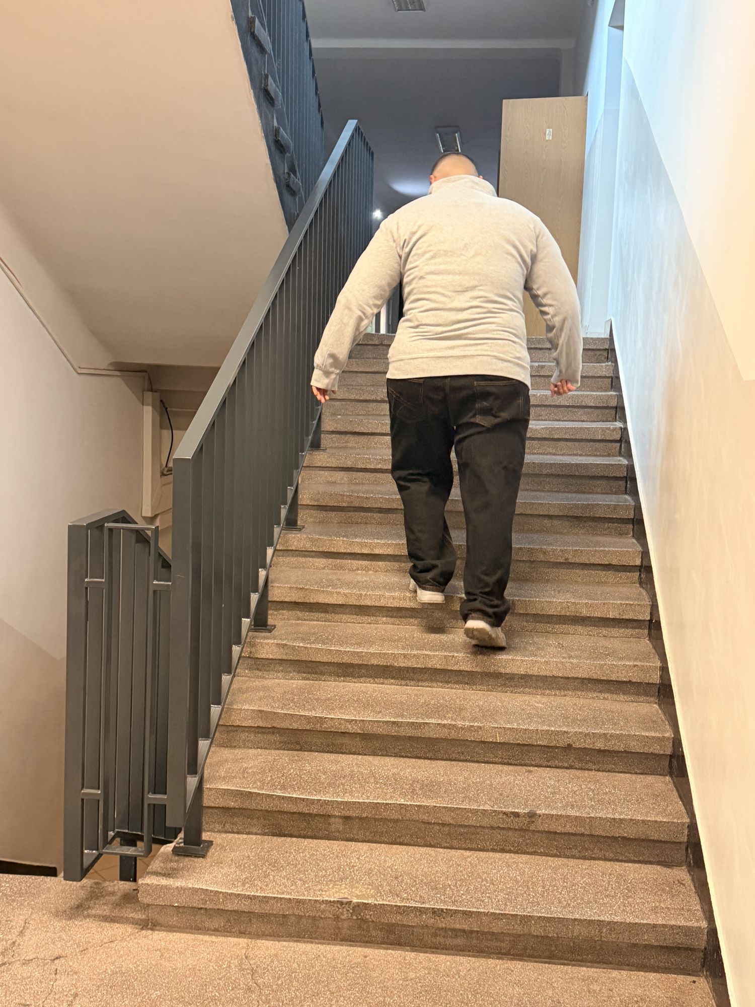 Uczestnik porusza się po schodach znajdujących się wewnątrz budynku. Zdjęcie przedstawia klatkę schodową z metalową balustradą oraz jasne, funkcjonalnie zaprojektowane wnętrze. Przestrzeń zapewnia wygodną komunikację pomiędzy kondygnacjami budynku.