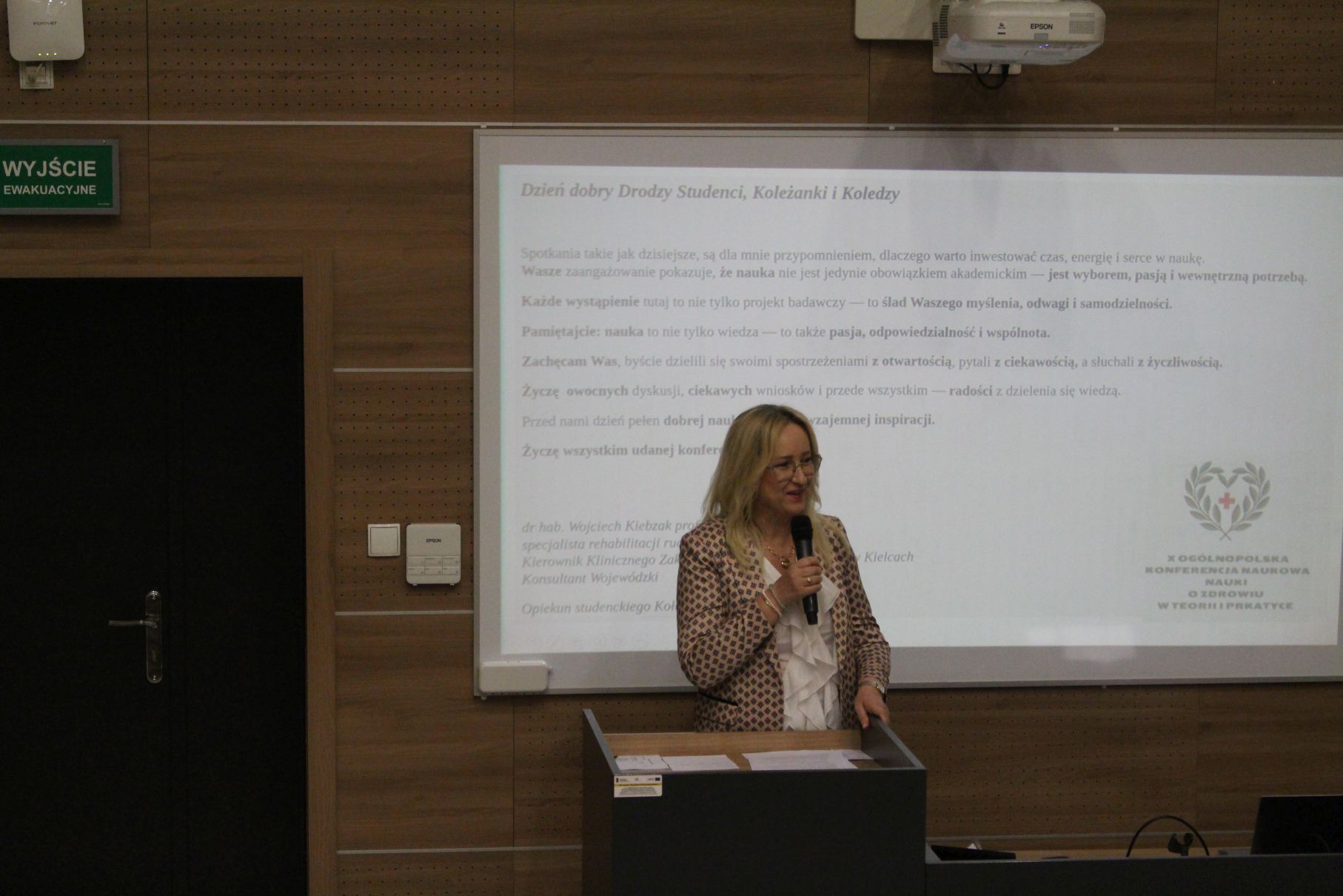 Prelegentka prowadzi wystąpienie przy mównicy podczas wydarzenia odbywającego się w sali konferencyjnej. W tle widoczny jest ekran z prezentacją zawierającą treści skierowane do uczestników spotkania. Zdjęcie przedstawia moment wykładu lub prezentacji edukacyjnej.
