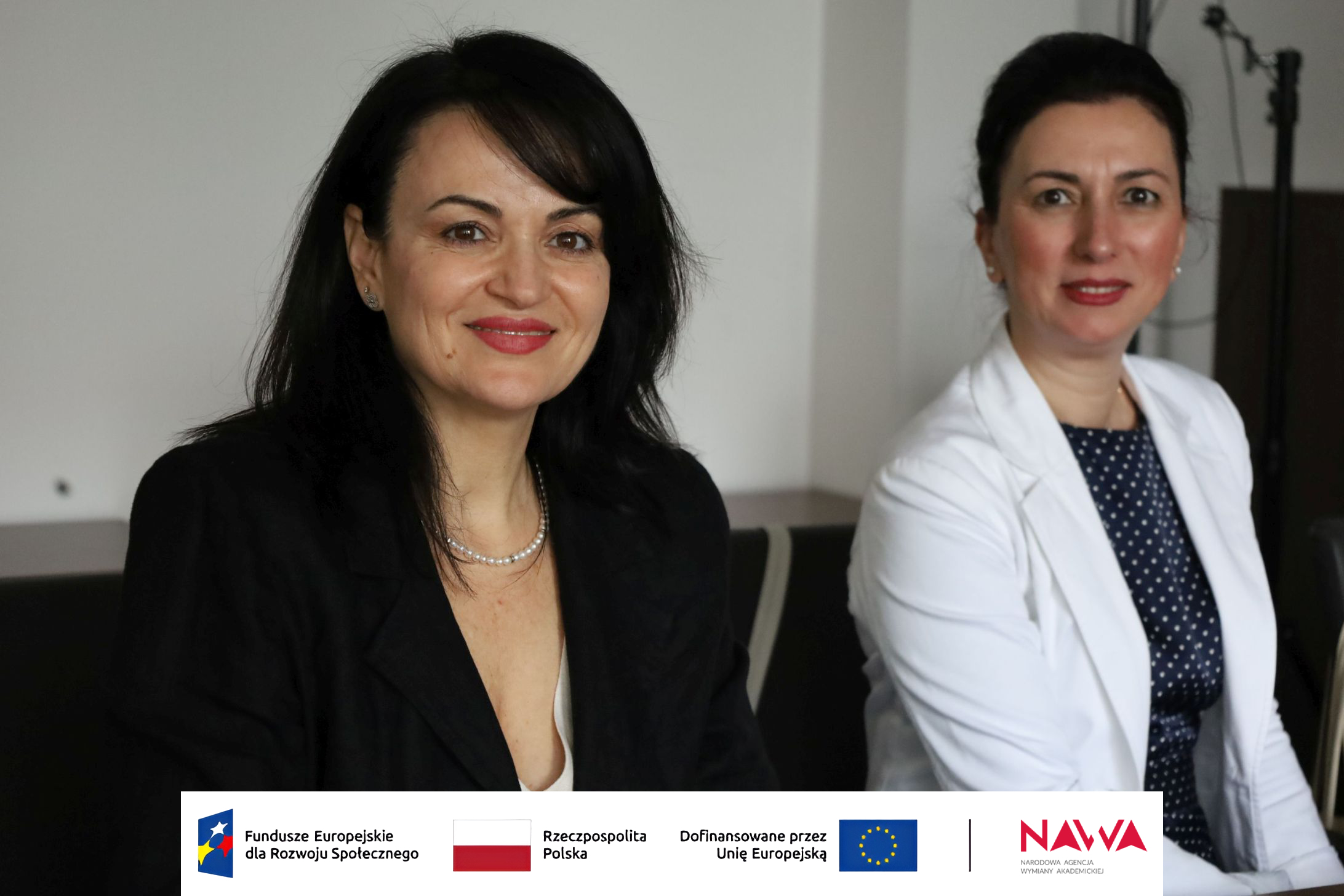 Dwie osoby siedzą obok siebie przy stole w pomieszczeniu i uśmiechają się do aparatu. W tle widoczne jest wyposażenie wnętrza, a rozmyte tło kieruje uwagę na osoby znajdujące się na pierwszym planie.