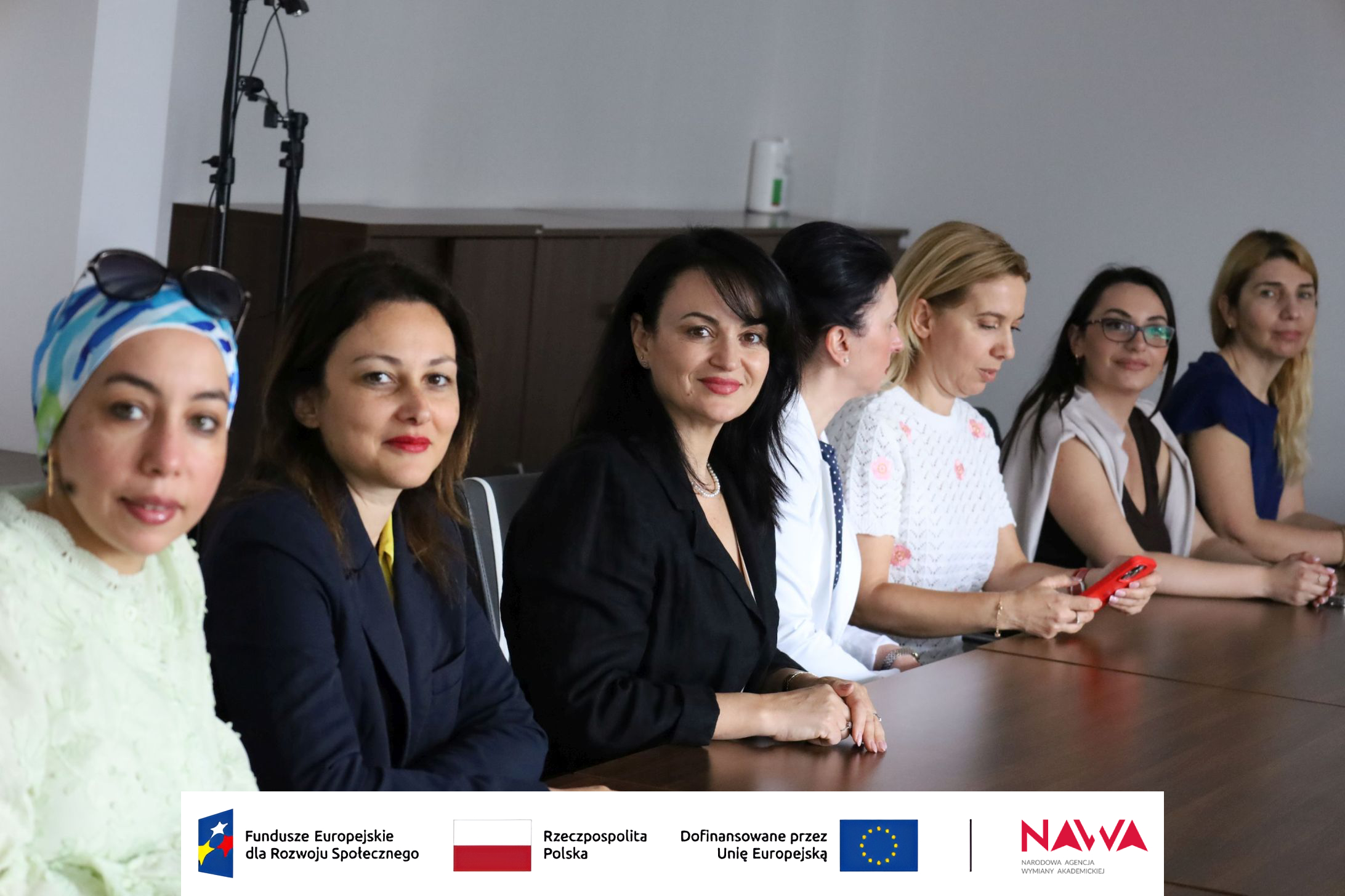Grupa osób siedzi przy stole konferencyjnym podczas spotkania w sali. Uczestniczki są skupione na rozmowie i materiałach znajdujących się na stole, a część z nich korzysta z telefonów. Zdjęcie przedstawia kameralne spotkanie w przestrzeni przeznaczonej do pracy i dyskusji.