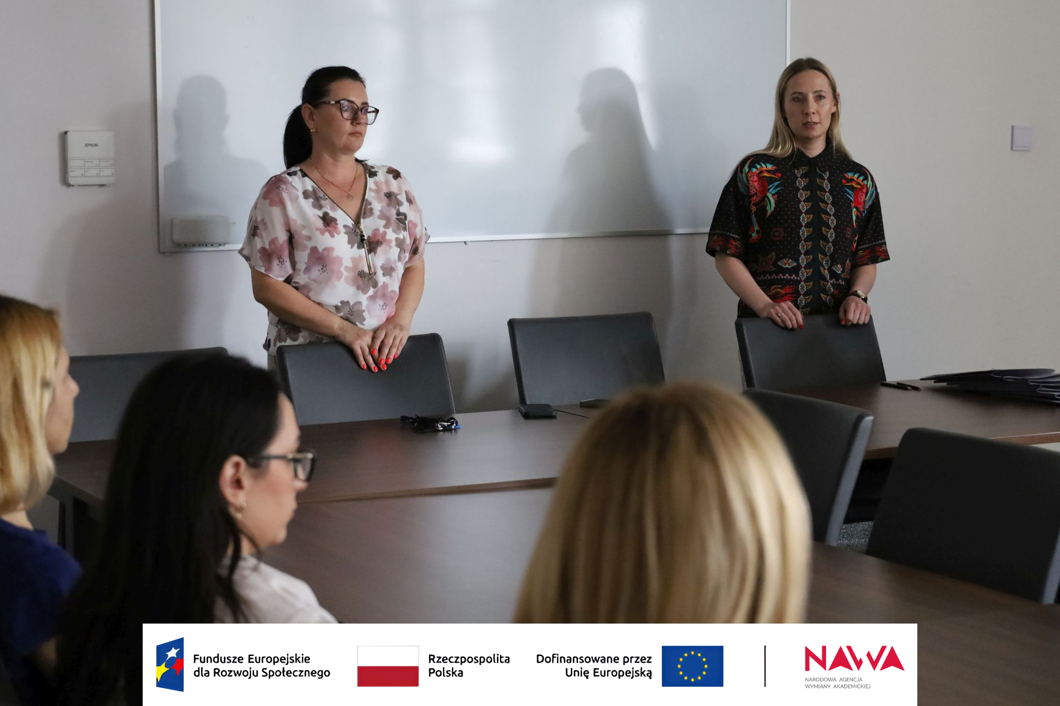 Spotkanie odbywające się w sali konferencyjnej z udziałem kilku osób. Część uczestniczek siedzi przy stole, a dwie osoby stoją na jego końcu, prowadząc lub uczestnicząc w dyskusji. W pomieszczeniu znajduje się stół konferencyjny, krzesła oraz tablica wykorzystywana podczas spotkań.