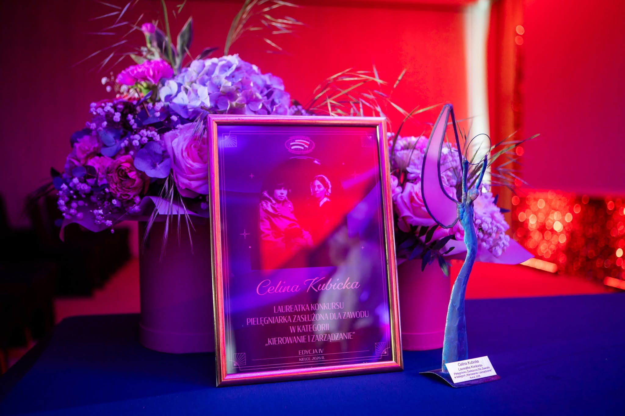 Na elegancko przygotowanym stole zaprezentowano statuetkę oraz oprawiony dyplom lub wyróżnienie związane z wydarzeniem o charakterze uroczystym. Kompozycję uzupełniają dekoracje kwiatowe oraz nastrojowe oświetlenie w odcieniach różu i fioletu, podkreślające prestiżowy charakter ceremonii. Całość sugeruje galę, konkurs, jubileusz lub inną okazję związaną z wręczaniem nagród i wyróżnień.