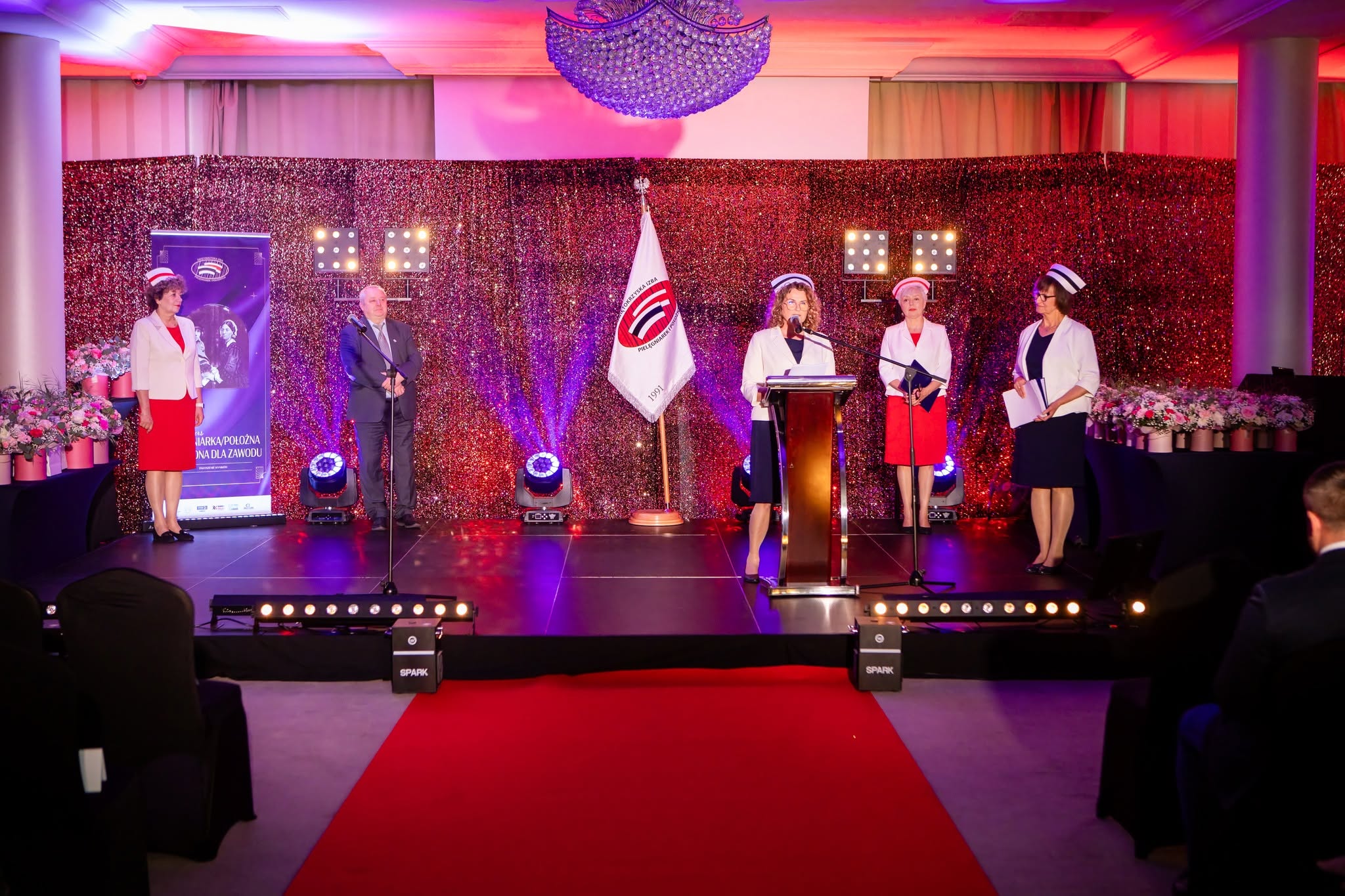 Na scenie odbywa się uroczysta gala lub ceremonia wręczenia wyróżnień. Osoba stojąca przy mównicy przemawia do zgromadzonej publiczności, a obok niej znajdują się pozostali uczestnicy wydarzenia pełniący funkcje organizacyjne lub reprezentacyjne. Efektowna oprawa sceniczna, dekoracje kwiatowe oraz profesjonalne oświetlenie podkreślają oficjalny i podniosły charakter uroczystości.