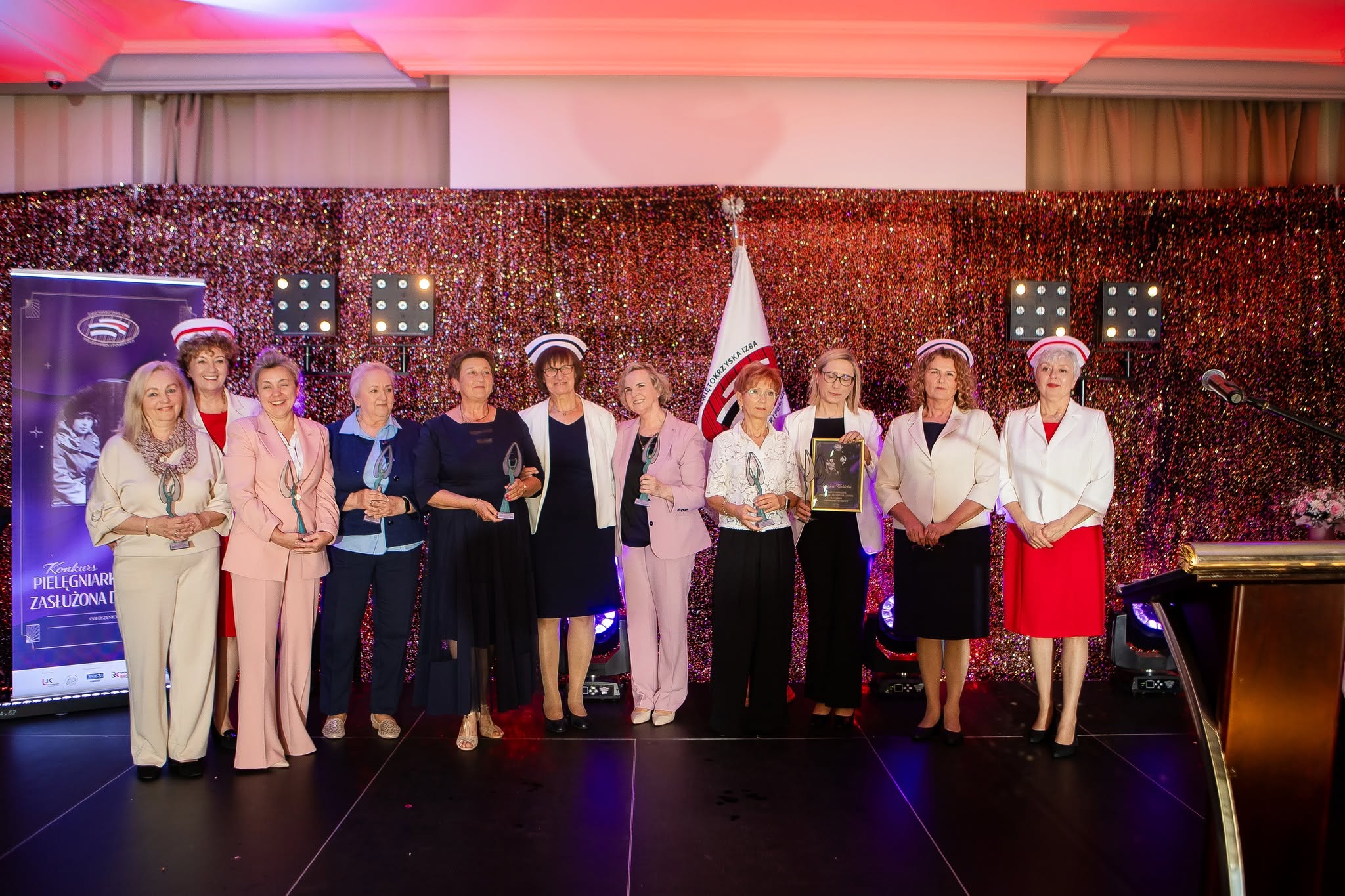 Grupa kobiet pozuje do pamiątkowego zdjęcia na scenie podczas uroczystej gali lub ceremonii wręczenia nagród. Uczestniczki trzymają statuetki i wyróżnienia, co wskazuje na uhonorowanie ich osiągnięć lub działalności zawodowej. Elegancka oprawa sceniczna, dekoracje oraz profesjonalne oświetlenie podkreślają prestiżowy i celebracyjny charakter wydarzenia.