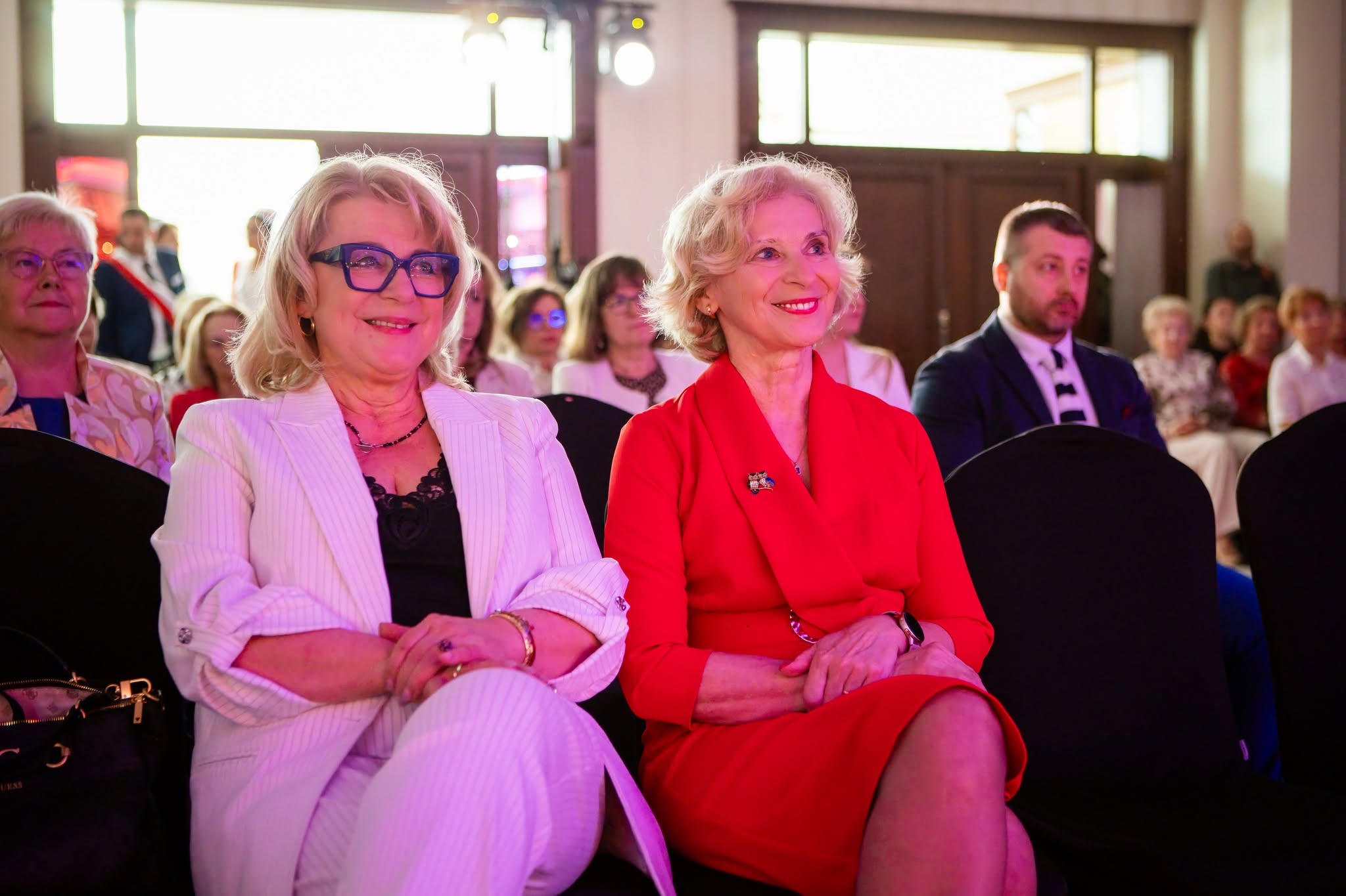 Uczestniczki wydarzenia siedzą na widowni i z zainteresowaniem obserwują przebieg uroczystości. W tle widoczni są pozostali goście zgromadzeni na sali, co podkreśla oficjalny charakter spotkania. Atmosfera jest przyjazna i podniosła, a zdjęcie dokumentuje moment uczestnictwa w gali, konferencji lub innym wydarzeniu okolicznościowym.