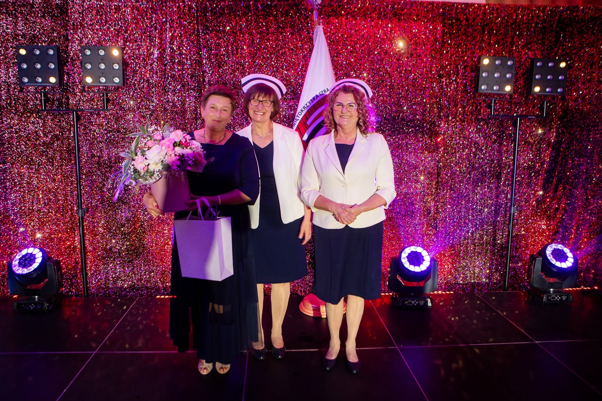 Trzy kobiety pozują do wspólnego zdjęcia na scenie podczas uroczystej gali lub ceremonii wręczenia wyróżnień. Jedna z uczestniczek trzyma bukiet kwiatów oraz upominek, co sugeruje moment podziękowania lub uhonorowania jej osiągnięć. Efektowne oświetlenie, dekoracyjne tło i oficjalna oprawa wydarzenia podkreślają podniosły oraz celebracyjny charakter uroczystości.