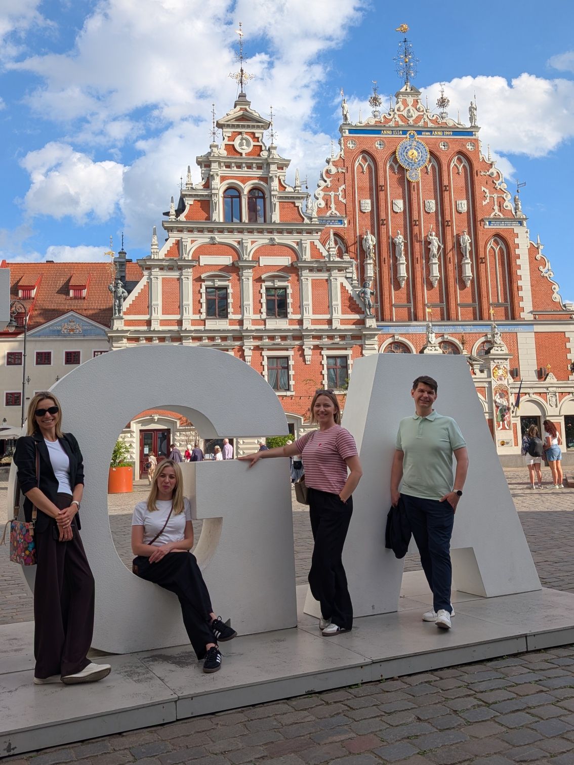 Grupa uczestników pozuje do pamiątkowego zdjęcia podczas wyjazdu lub wizyty studyjnej w historycznej części miasta. W tle widoczne są zabytkowe budynki o bogato zdobionej architekturze, stanowiące charakterystyczny element miejskiego krajobrazu. Słoneczna pogoda i swobodna atmosfera podkreślają rekreacyjny oraz integracyjny charakter wspólnego wyjazdu.