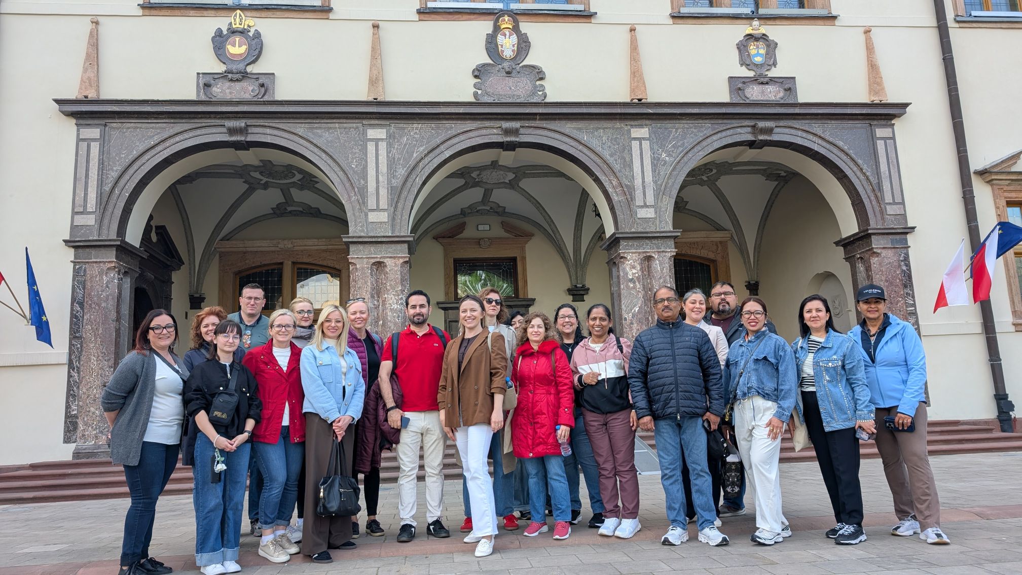 Grupa uczestników pozuje do wspólnego zdjęcia przed reprezentacyjnym wejściem do zabytkowego budynku o bogato zdobionej architekturze. Charakterystyczne arkady, kolumny oraz herby nad wejściami podkreślają historyczny i prestiżowy charakter obiektu. Uczestnicy stoją razem w przyjaznej atmosferze, dokumentując wspólne wydarzenie lub wizytę.