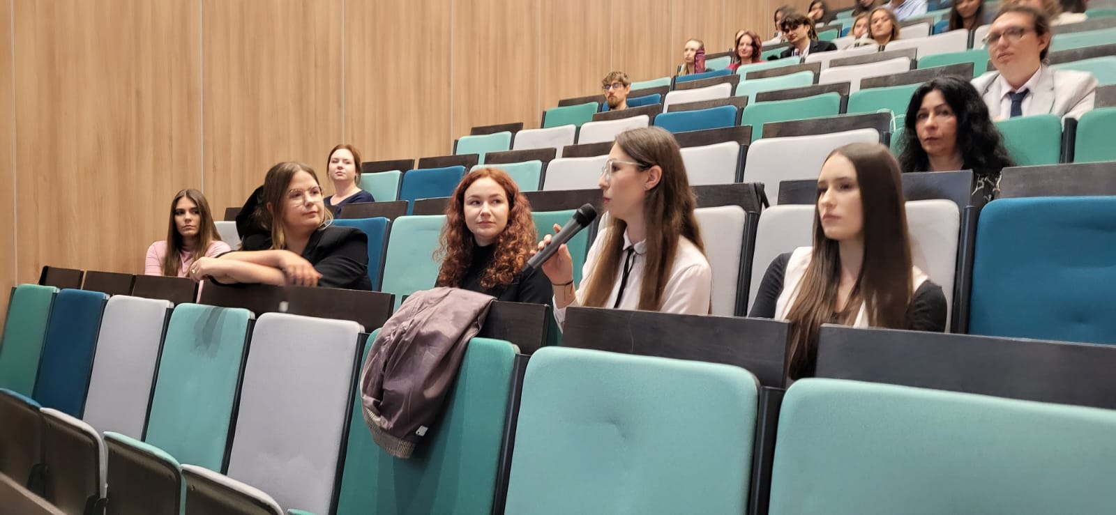 Uczestniczka wydarzenia zabiera głos podczas sesji dyskusyjnej lub panelu pytań i odpowiedzi w auli wykładowej. Pozostali uczestnicy siedzą na widowni i z uwagą śledzą przebieg rozmowy. Nowoczesna sala z amfiteatralnym układem miejsc sprzyja organizacji konferencji, seminariów i spotkań naukowych.