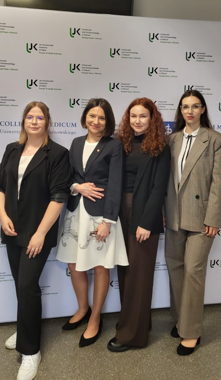 Cztery osoby pozują do pamiątkowego zdjęcia na tle ścianki promocyjnej Uniwersytetu Jana Kochanowskiego w Kielcach. Tło zawiera logotypy oraz nazwę jednostki związanej z naukami o zdrowiu, co wskazuje na udział w wydarzeniu akademickim, konferencji lub spotkaniu naukowym. Uczestnicy ubrani są w strój formalny i półformalny, podkreślający oficjalny charakter okazji.