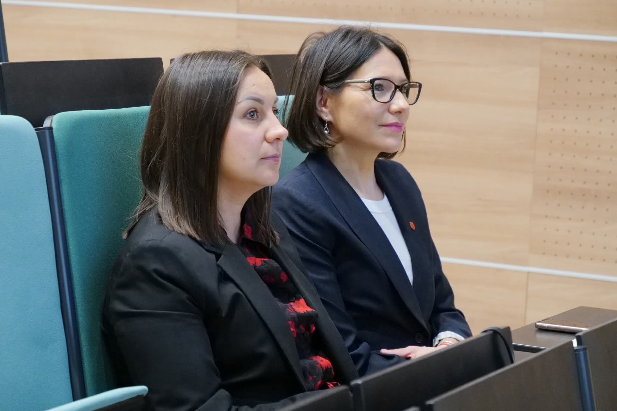 Dwie uczestniczki konferencji lub seminarium siedzą na widowni i przysłuchują się prezentacji lub dyskusji. Nowoczesne fotele oraz drewniane wykończenie ścian tworzą profesjonalne otoczenie charakterystyczne dla auli wykładowej lub sali konferencyjnej. Atmosfera wydarzenia sprzyja skupieniu i aktywnemu uczestnictwu w spotkaniu naukowym lub edukacyjnym.