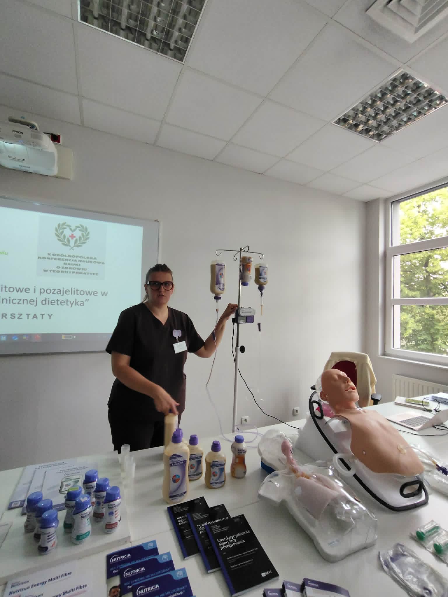 W sali szkoleniowej prowadzona jest prezentacja dotycząca żywienia klinicznego lub opieki medycznej. Na stanowisku demonstracyjnym znajdują się preparaty żywieniowe, materiały edukacyjne oraz fantom pacjenta wykorzystywany do ćwiczeń praktycznych. Prowadząca omawia działanie sprzętu i procedur przy stojaku z workami infuzyjnymi, a prezentacja multimedialna wspiera przekazywane treści.