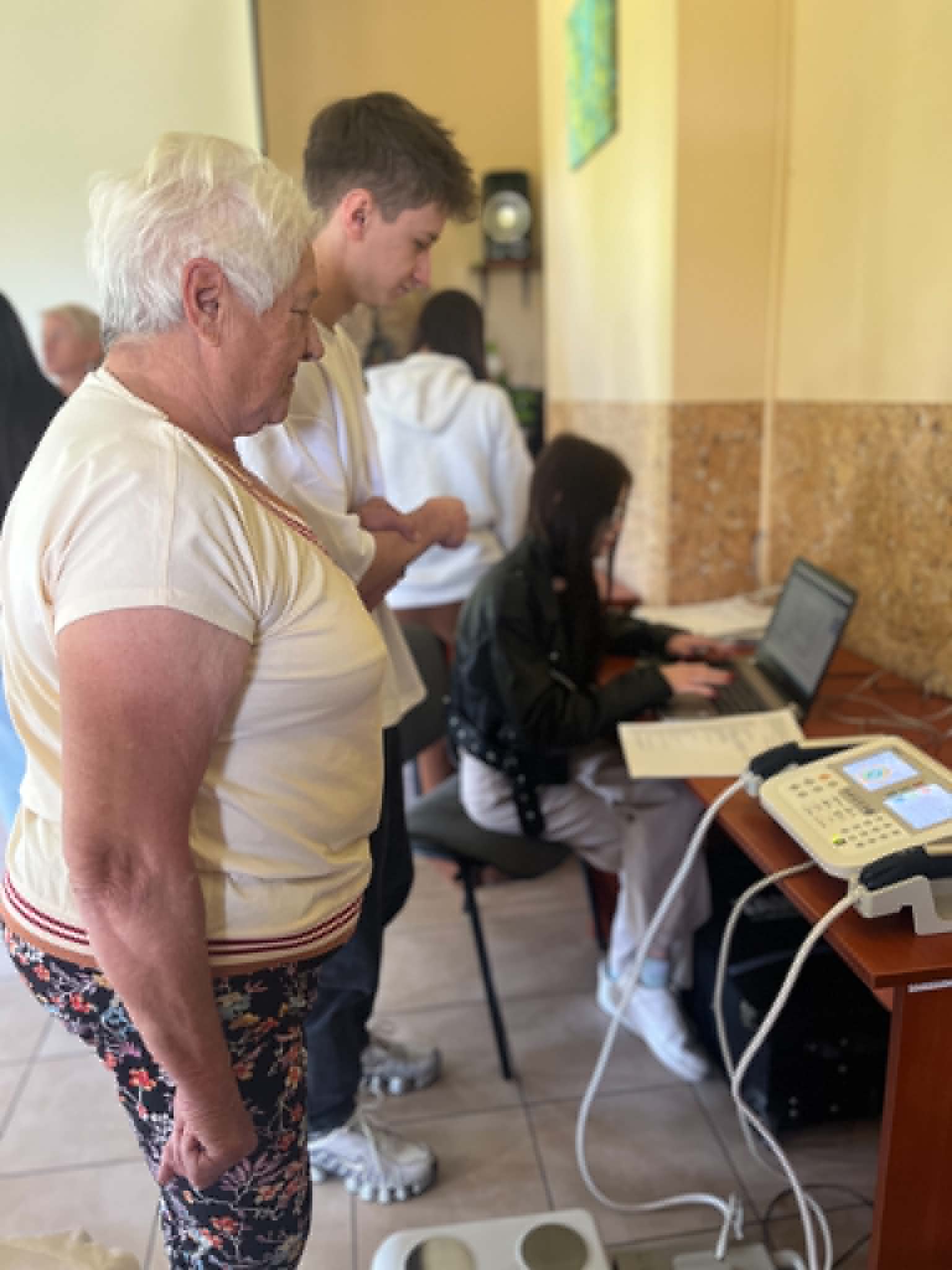 Uczestniczka bierze udział w badaniu lub konsultacji zdrowotnej przy wykorzystaniu specjalistycznego urządzenia pomiarowego. Obok znajduje się osoba nadzorująca przebieg pomiaru, natomiast w tle widoczna jest osoba dokumentująca wyniki na laptopie i w formularzach. Całość odbywa się w pomieszczeniu o charakterze profilaktycznym lub diagnostycznym.