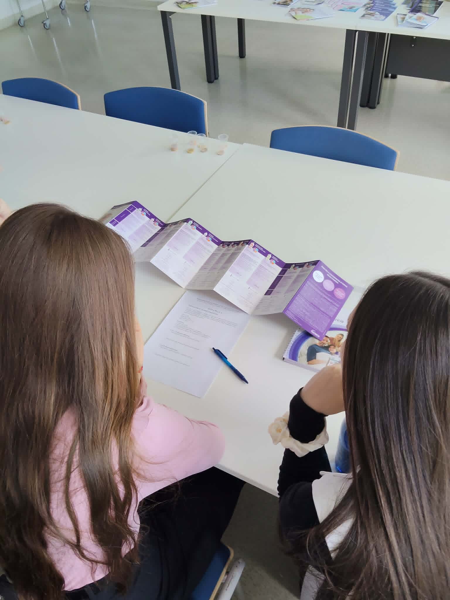 Dwie uczestniczki zajęć siedzą przy stole i zapoznają się z materiałami edukacyjnymi w formie broszur lub ulotek informacyjnych. Na blacie znajdują się również notatki oraz przybory do pisania, wykorzystywane podczas szkolenia lub warsztatów. Jasne wnętrze i przygotowane materiały wskazują na edukacyjny charakter spotkania.