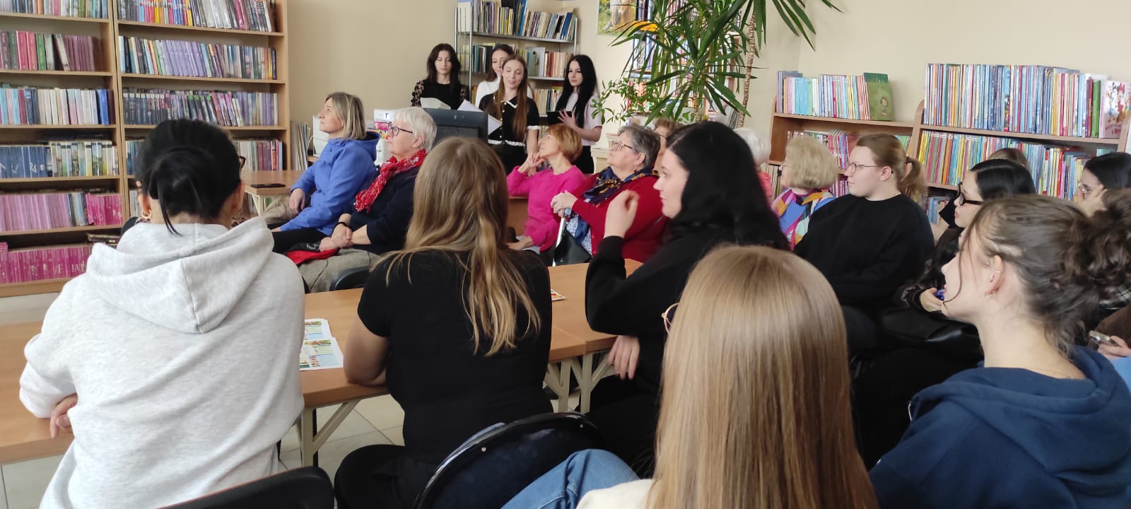 Uczestnicy spotkania siedzą w bibliotece wśród regałów wypełnionych książkami i słuchają prezentacji lub wykładu prowadzonego przez osoby stojące z przodu sali. W wydarzeniu biorą udział osoby w różnym wieku, a atmosfera jest skupiona i sprzyja zdobywaniu wiedzy. Wnętrze biblioteki oraz obecność materiałów edukacyjnych podkreślają popularyzatorski i edukacyjny charakter spotkania.