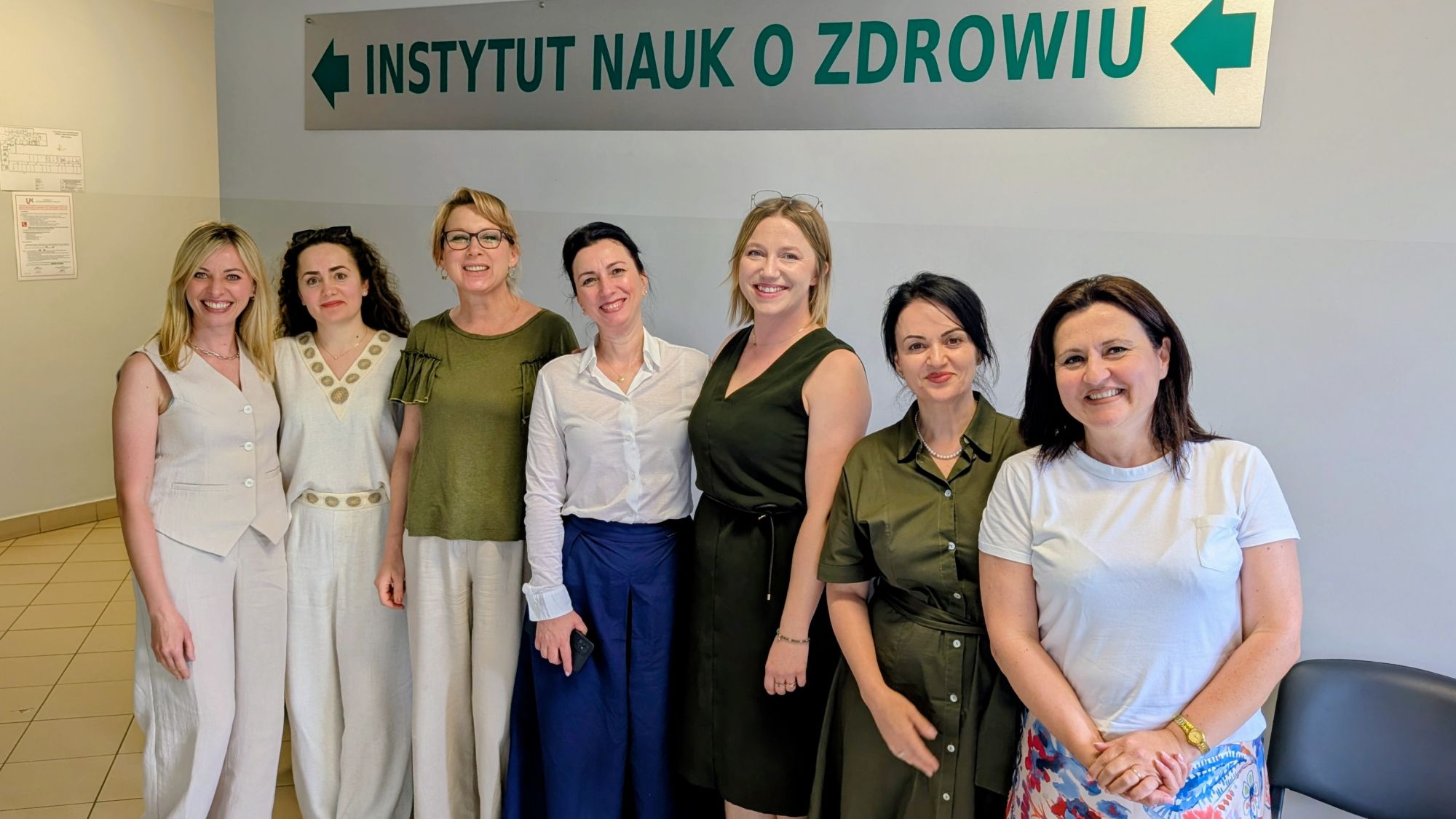 Zdjęcie przedstawia grupę uczestniczek pozujących do wspólnego zdjęcia w holu budynku. W tle widoczny jest znak kierujący do Instytutu Nauk o Zdrowiu. Fotografia została wykonana w przestrzeni uczelni i przedstawia uczestniczki wydarzenia lub spotkania.
