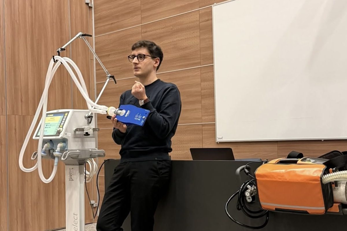 Zdjęcie przedstawia prowadzącego prezentującego specjalistyczny sprzęt medyczny podczas zajęć dydaktycznych. W sali widoczne są respirator oraz wyposażenie wykorzystywane do nauki procedur medycznych i ratunkowych. Fotografia ukazuje praktyczną część szkolenia z wykorzystaniem nowoczesnej aparatury medycznej.