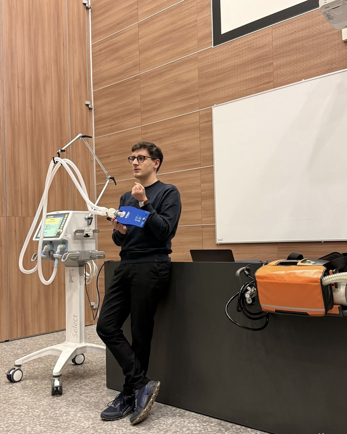 Zdjęcie przedstawia prowadzącego prezentującego specjalistyczny sprzęt medyczny podczas zajęć dydaktycznych. W sali widoczne są respirator oraz wyposażenie wykorzystywane do nauki procedur medycznych i ratunkowych. Fotografia ukazuje praktyczną część szkolenia z wykorzystaniem nowoczesnej aparatury medycznej.