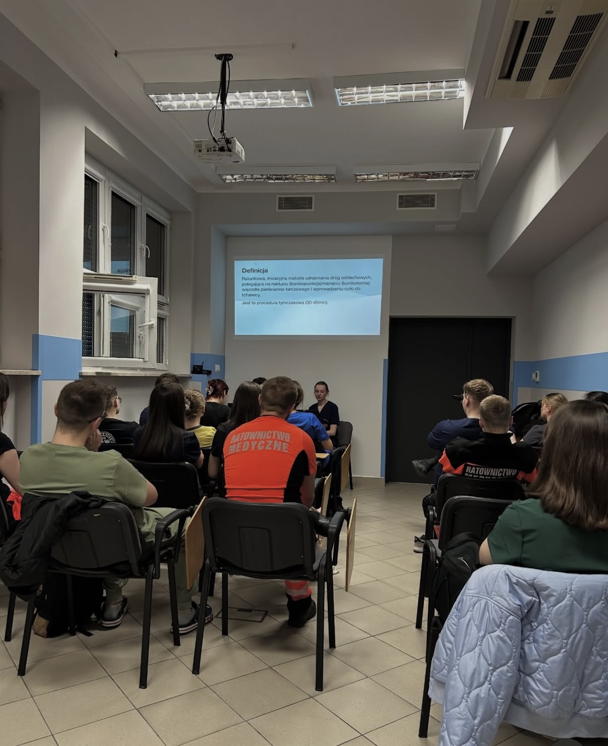 Zdjęcie przedstawia uczestnik&oacute;w podczas wykładu lub prezentacji w sali dydaktycznej. W tle widoczny jest ekran z wyświetlaną prezentacją wykorzystywaną podczas zajęć. Fotografia ukazuje teoretyczną część kształcenia z zakresu nauk medycznych i ratownictwa.