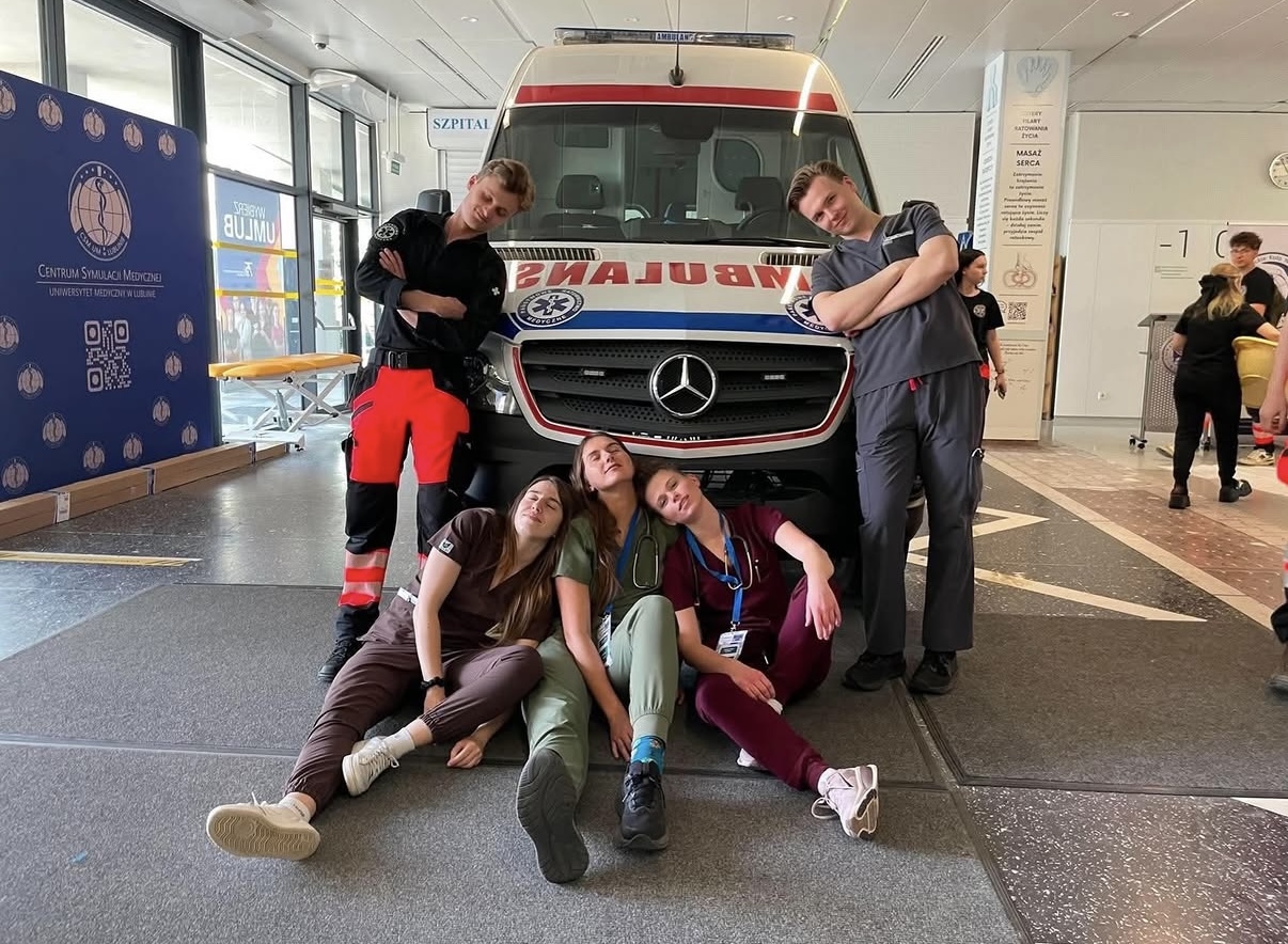 Zdjęcie przedstawia grupę uczestnik&oacute;w pozujących do wsp&oacute;lnego zdjęcia przed ambulansem. Fotografia została wykonana podczas wydarzenia lub szkolenia związanego z ratownictwem medycznym. W tle widoczny jest ambulans oraz elementy ekspozycji poświęconej medycynie ratunkowej.
