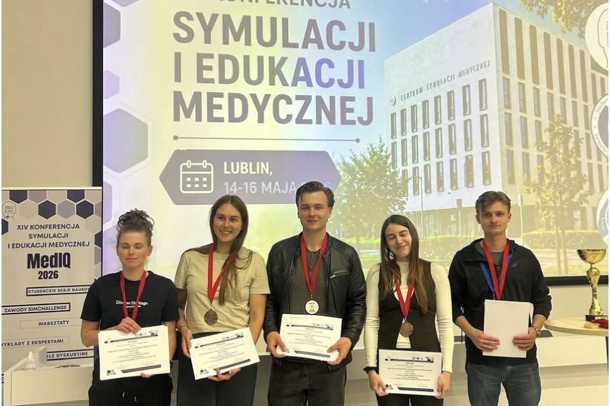 Zdjęcie przedstawia grupę uczestników pozujących do wspólnego zdjęcia podczas konferencji poświęconej symulacji i edukacji medycznej. Uczestnicy trzymają certyfikaty oraz medale, podkreślające udział i osiągnięcia podczas wydarzenia. W tle widoczny jest ekran z nazwą konferencji i informacjami o wydarzeniu.
