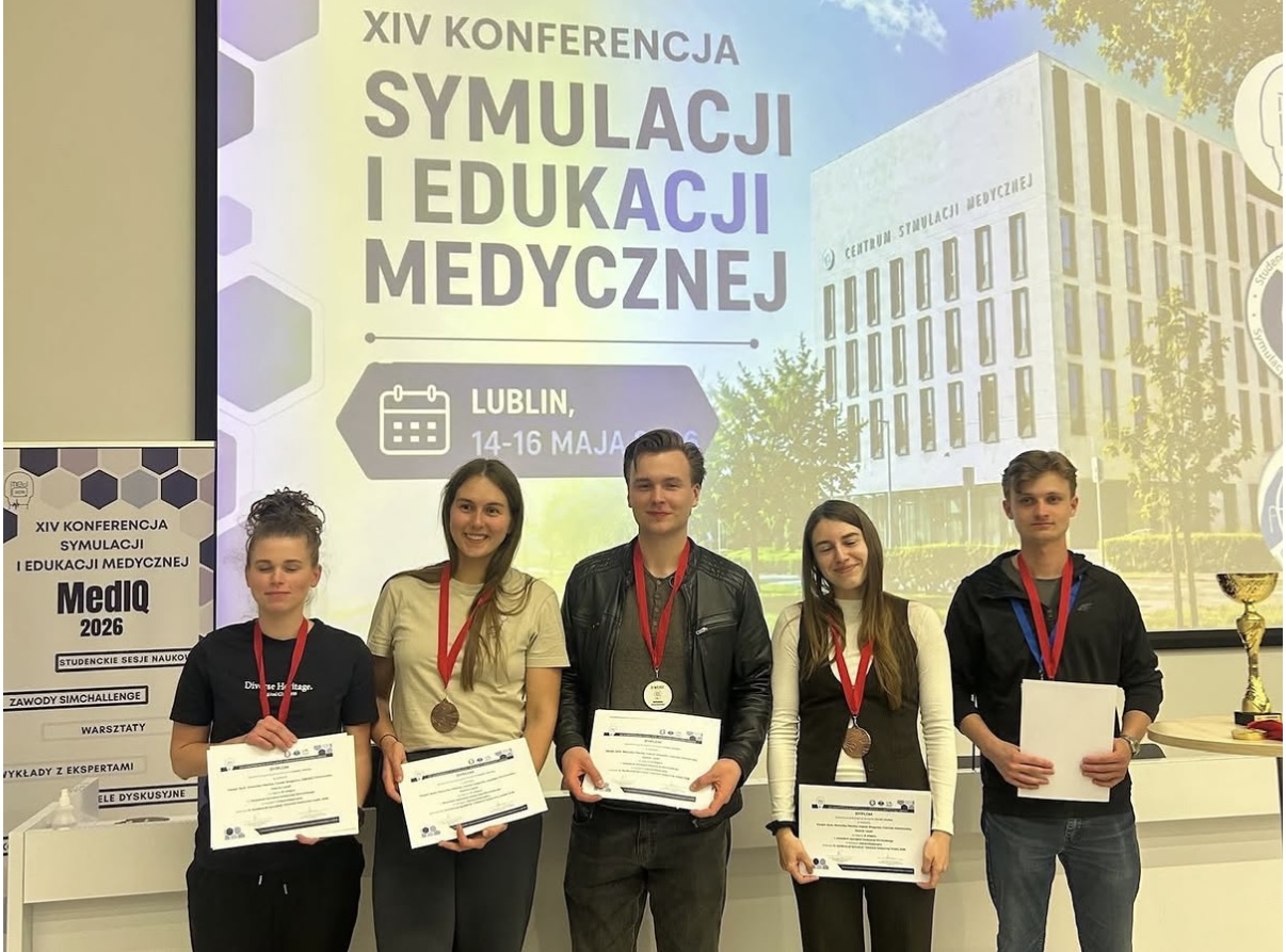 Zdjęcie przedstawia grupę uczestnik&oacute;w pozujących do wsp&oacute;lnego zdjęcia podczas konferencji poświęconej symulacji i edukacji medycznej. Uczestnicy trzymają certyfikaty oraz medale, podkreślające udział i osiągnięcia podczas wydarzenia. W tle widoczny jest ekran z nazwą konferencji i informacjami o wydarzeniu.