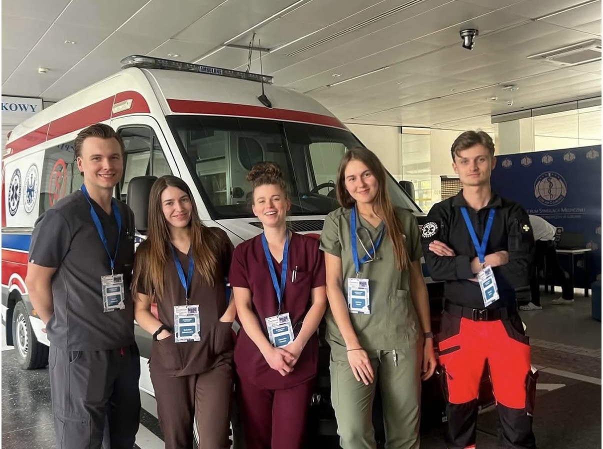 Zdjęcie przedstawia grupę uczestnik&oacute;w pozujących do wsp&oacute;lnego zdjęcia przed ambulansem. Fotografia została wykonana podczas wydarzenia lub szkolenia związanego z ratownictwem medycznym. W tle widoczny jest ambulans oraz elementy ekspozycji poświęconej medycynie ratunkowej.