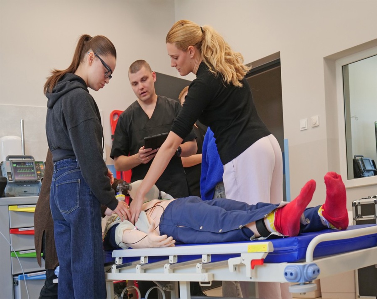 Zdjęcie przedstawia ćwiczenia praktyczne prowadzone w sali symulacji medycznej z wykorzystaniem fantomu szkoleniowego. Uczestnicy wykonują procedury medyczne pod nadzorem prowadzącego, korzystając ze specjalistycznego sprzętu i wyposażenia. Fotografia ukazuje praktyczną naukę umiejętności klinicznych w warunkach zbliżonych do rzeczywistych.