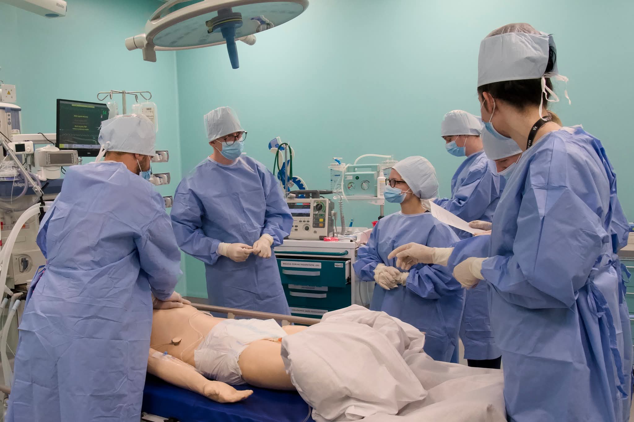 Zdjęcie przedstawia ćwiczenia praktyczne prowadzone w sali symulacji medycznej. Uczestnicy wykonują procedury medyczne z wykorzystaniem fantomu oraz specjalistycznego sprzętu. Pomieszczenie odzwierciedla warunki bloku operacyjnego lub sali zabiegowej, umożliwiając naukę praktycznych umiejętności.