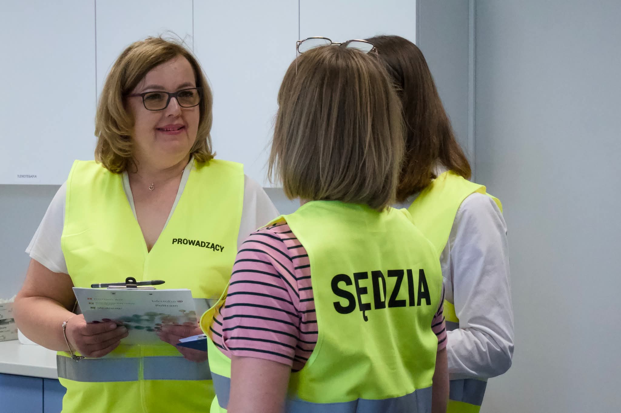 Zdjęcie przedstawia trzy osoby rozmawiające w nowoczesnym pomieszczeniu dydaktycznym. Uczestnicy mają na sobie kamizelki z oznaczeniami pełnionych r&oacute;l i omawiają dokumenty. W tle widoczne są szafki oraz wyposażenie sali, podkreślające profesjonalny charakter wydarzenia.